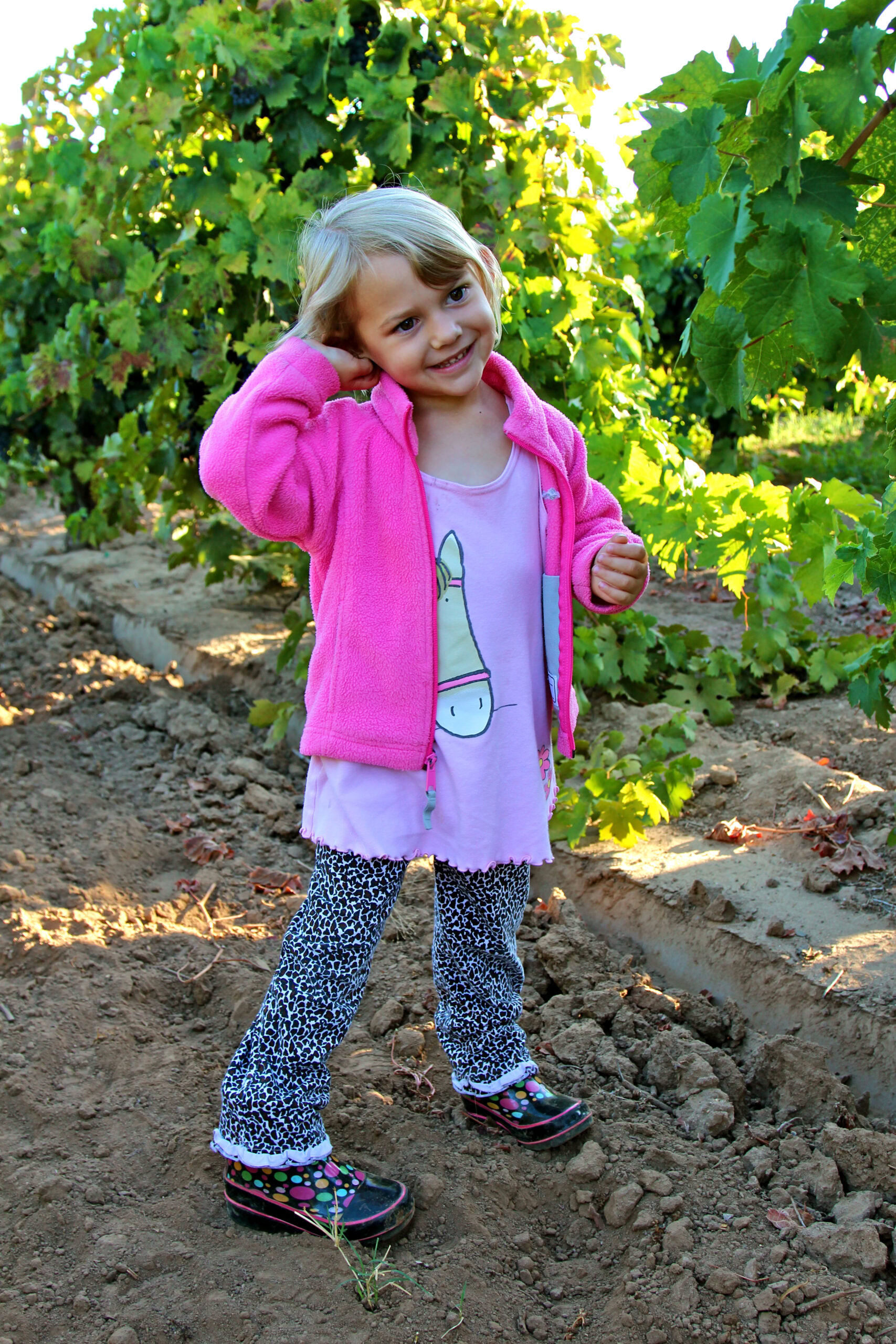Young girl smiling in a vineyard, wearing a pink jacket and patterned pants, enjoying a sunny day among grapevines.
