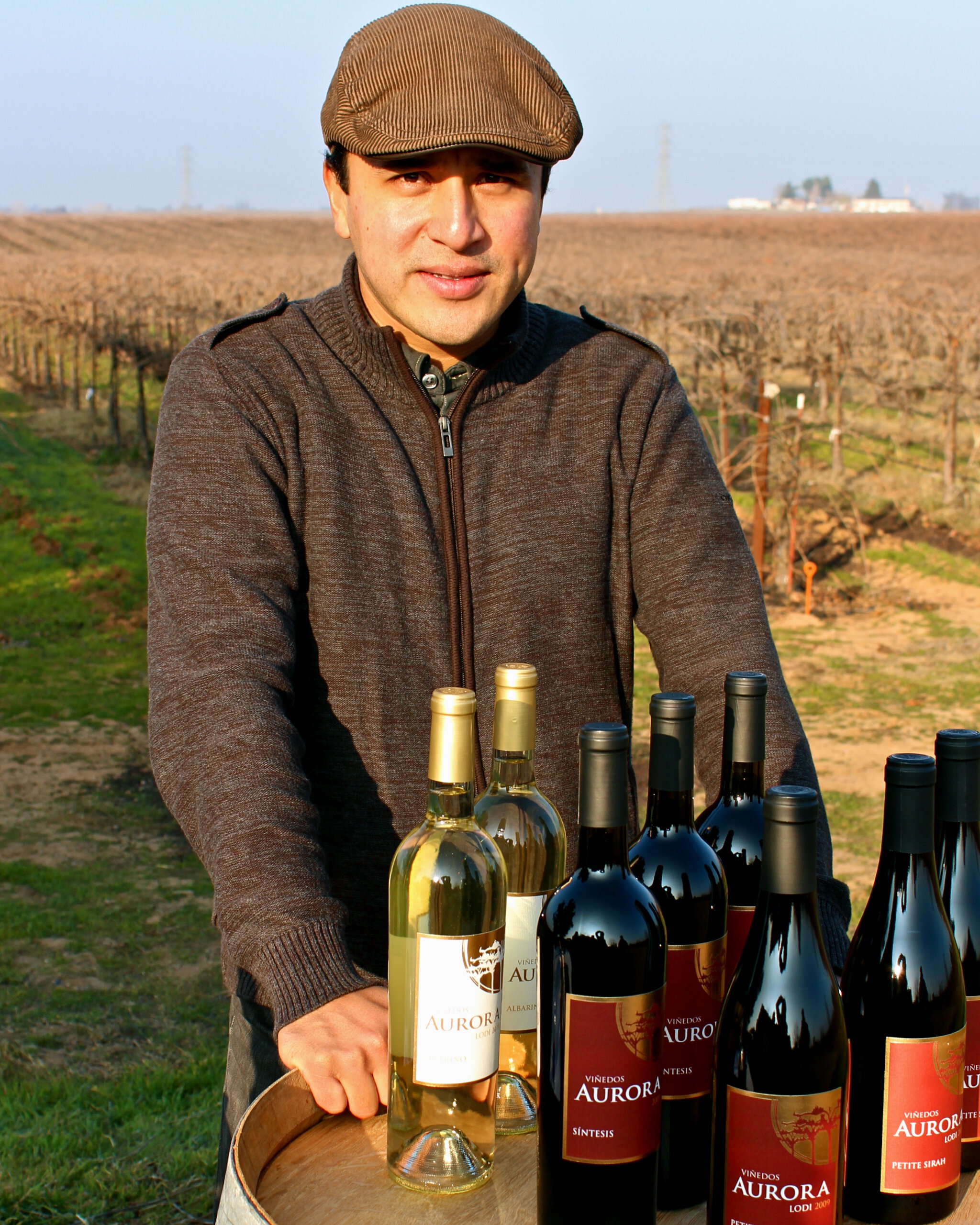 Man in vineyard with bottles of wine from Lodi Winegrape Commission, showcasing local wine production.