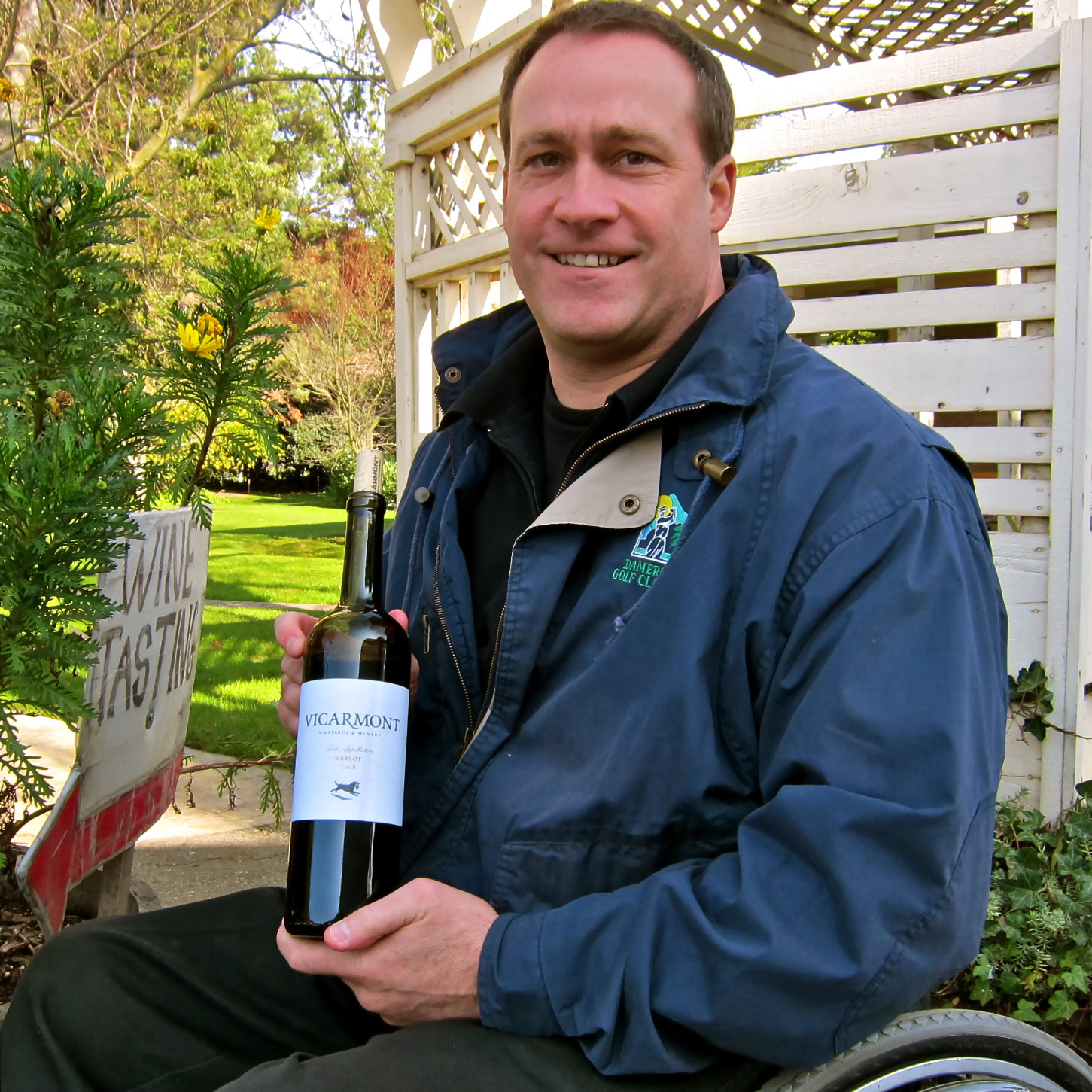 Man in a wheelchair holding a bottle of wine outdoors at a vineyard event.