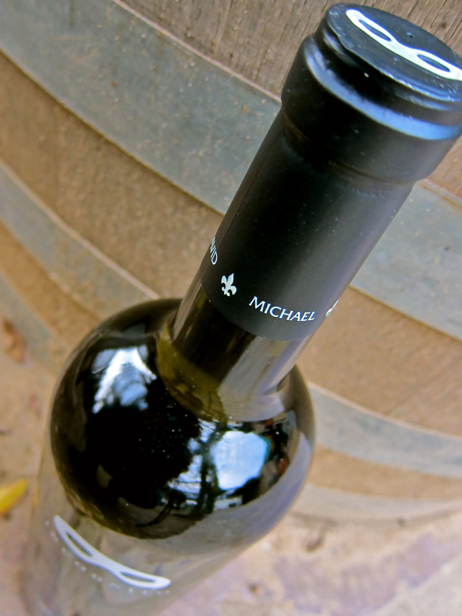 Close-up of a wine bottle with a black cap and label, set against a wooden barrel background.