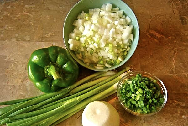 Fresh chopped onions, green bell pepper, scallions, and a white onion on a wooden surface.