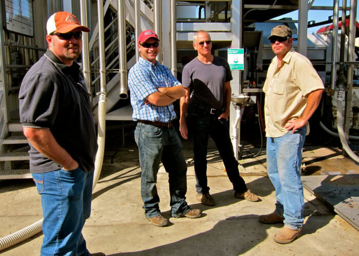 Group of four men standing outdoors at a winery, smiling, with wine production equipment in the background.