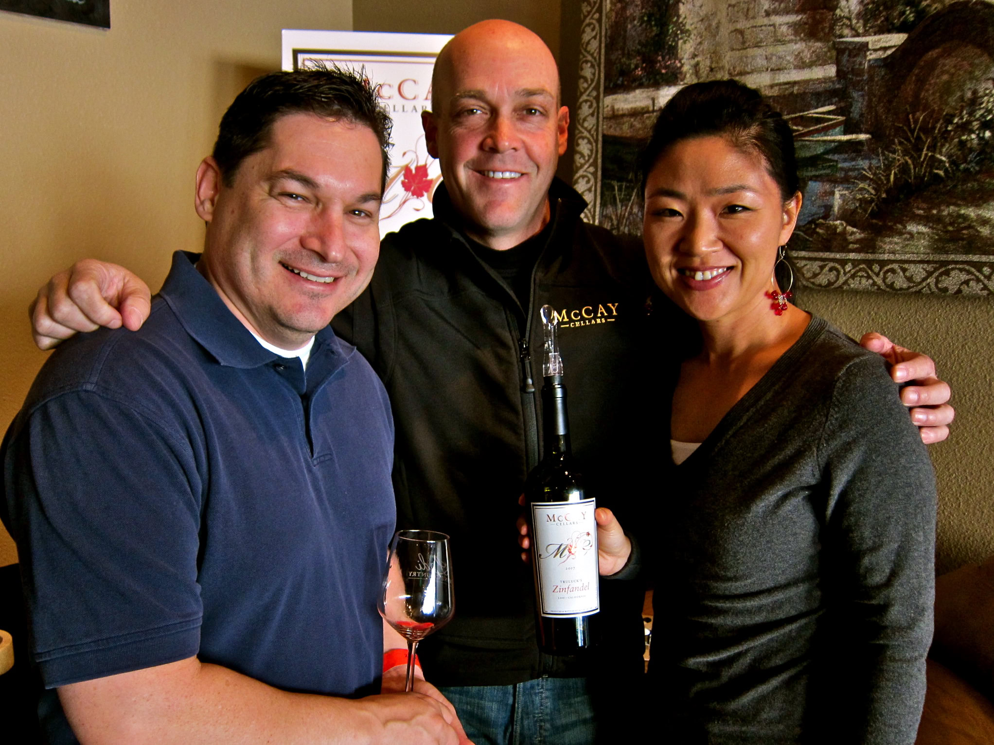 Group of three people at a wine tasting event, holding a bottle of wine, smiling, with a wine glass in hand.