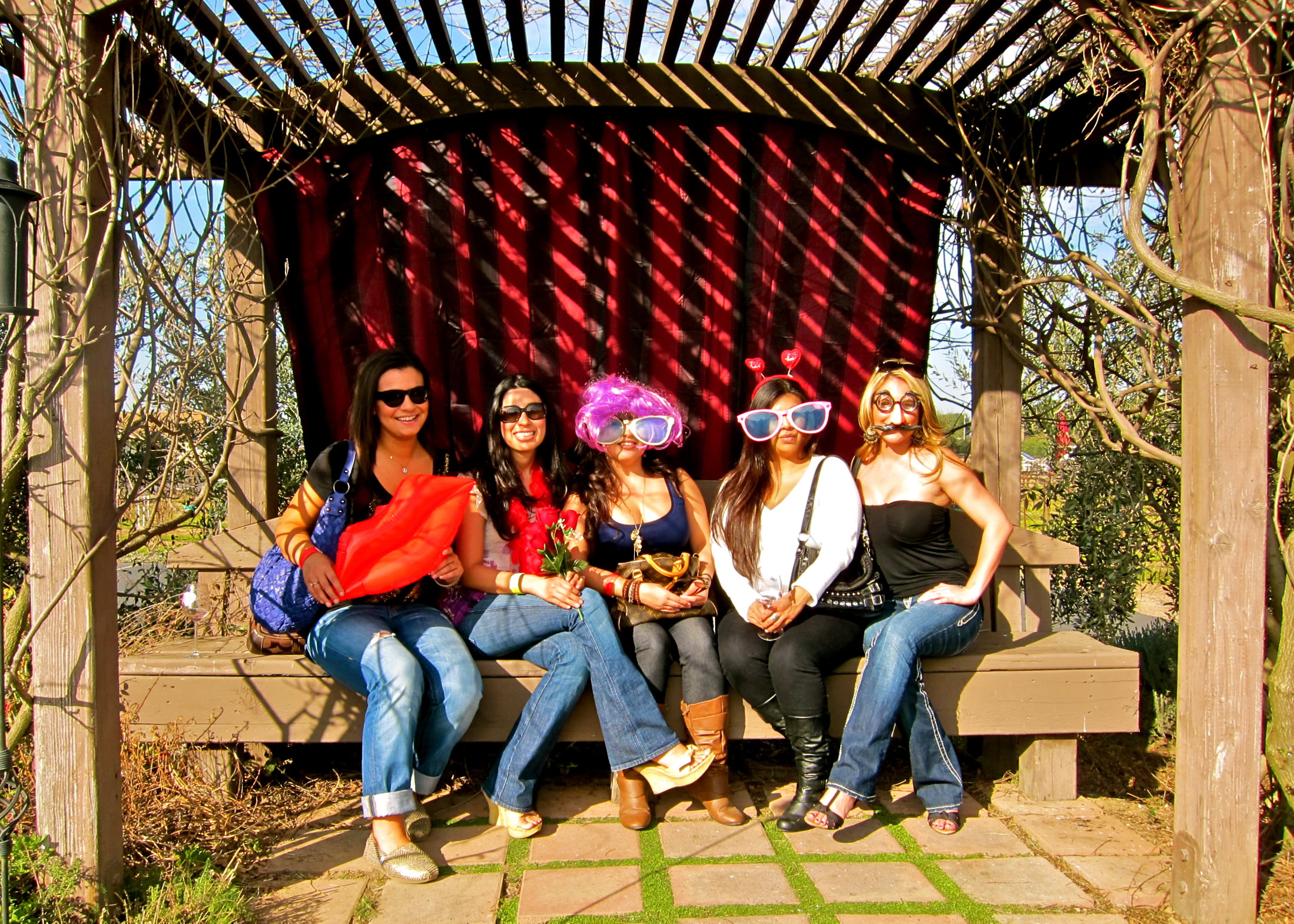 Women wearing fun accessories sitting on a wooden bench in a garden setting.