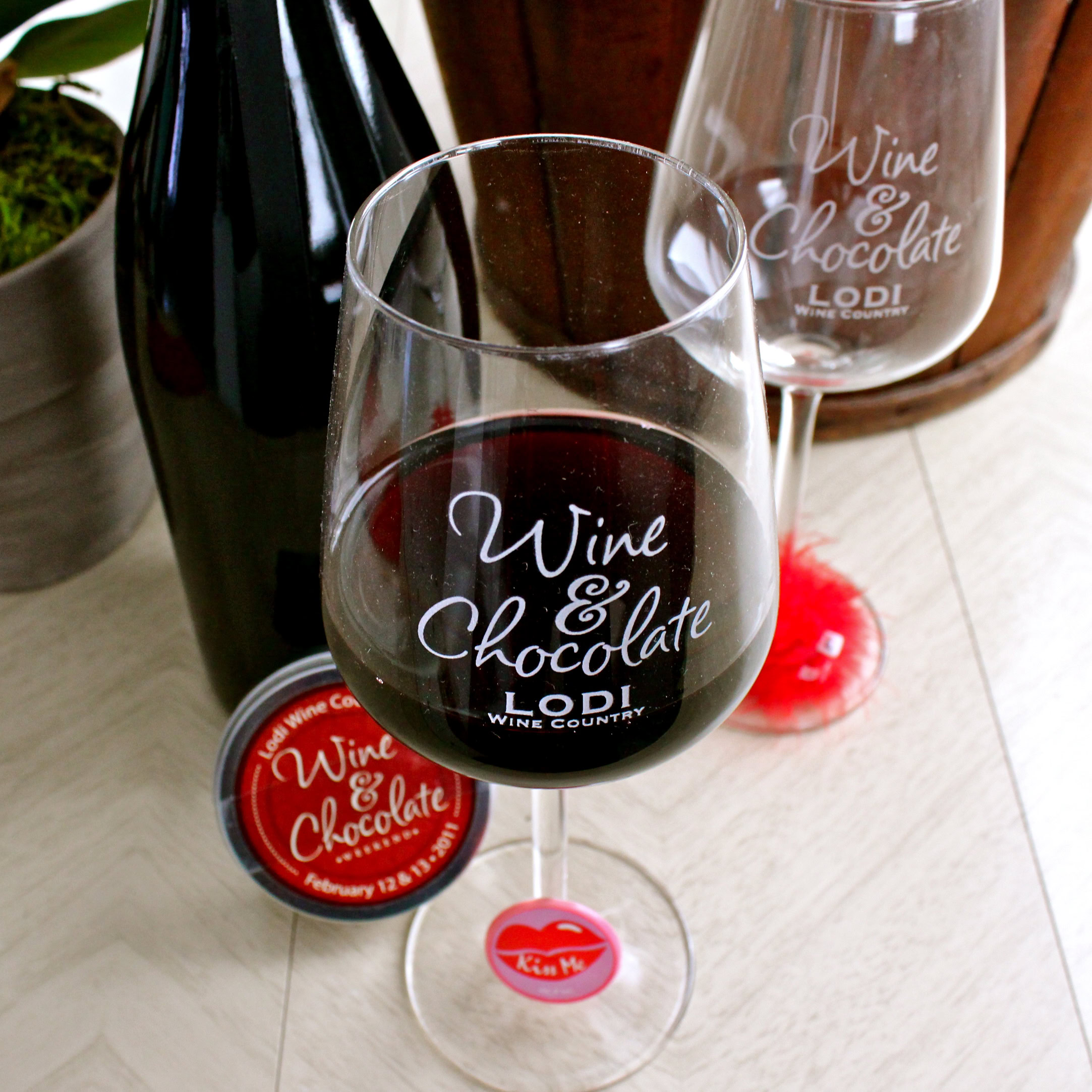 Glass of red wine with Lodi Wine Country branding, wine bottle, and chocolate on a wooden table.