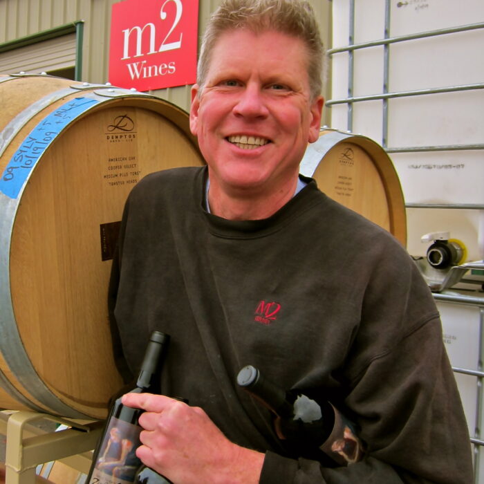 Winemaker holding a wine bottle in a winery with wine barrels in the background.