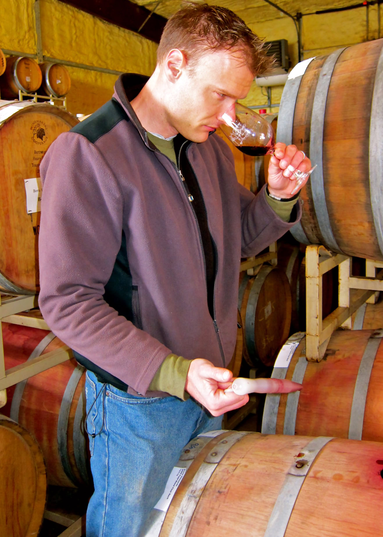 Man tasting wine from a glass in a winery with wine barrels in the background.
