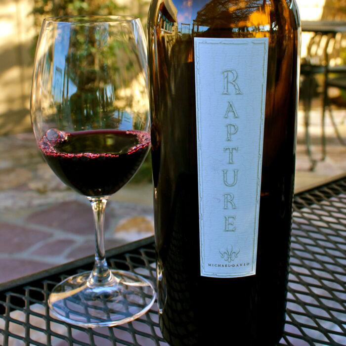 A wine bottle labeled 'Rapture' next to a glass of red wine on an outdoor table.