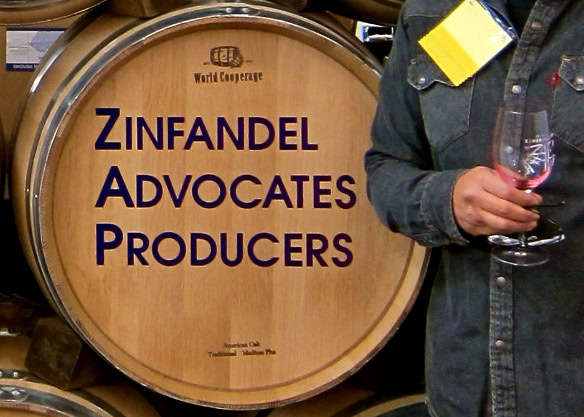 Person holding a glass of wine next to a barrel labeled Zinfandel Advocates Producers.