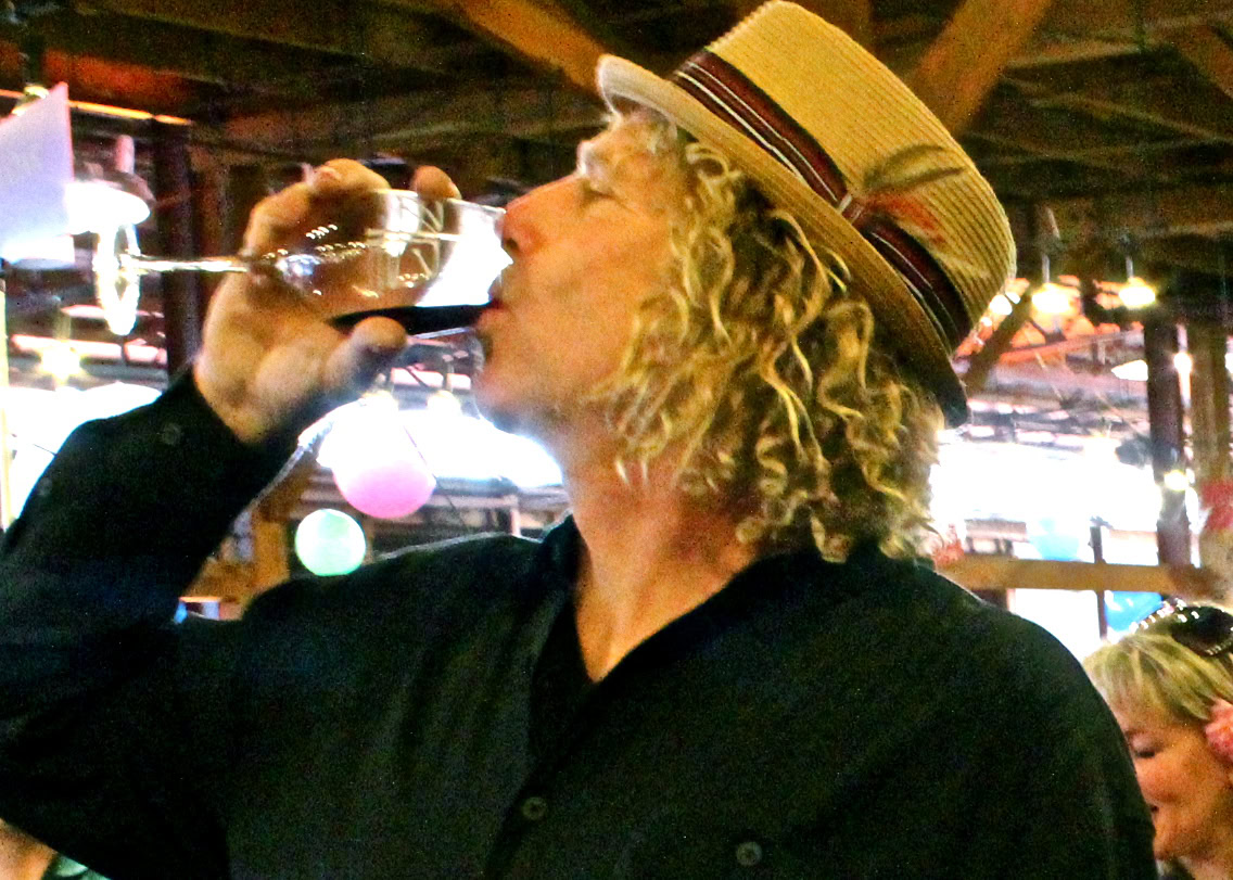 Woman with curly hair wearing a hat drinking wine at a wine event.
