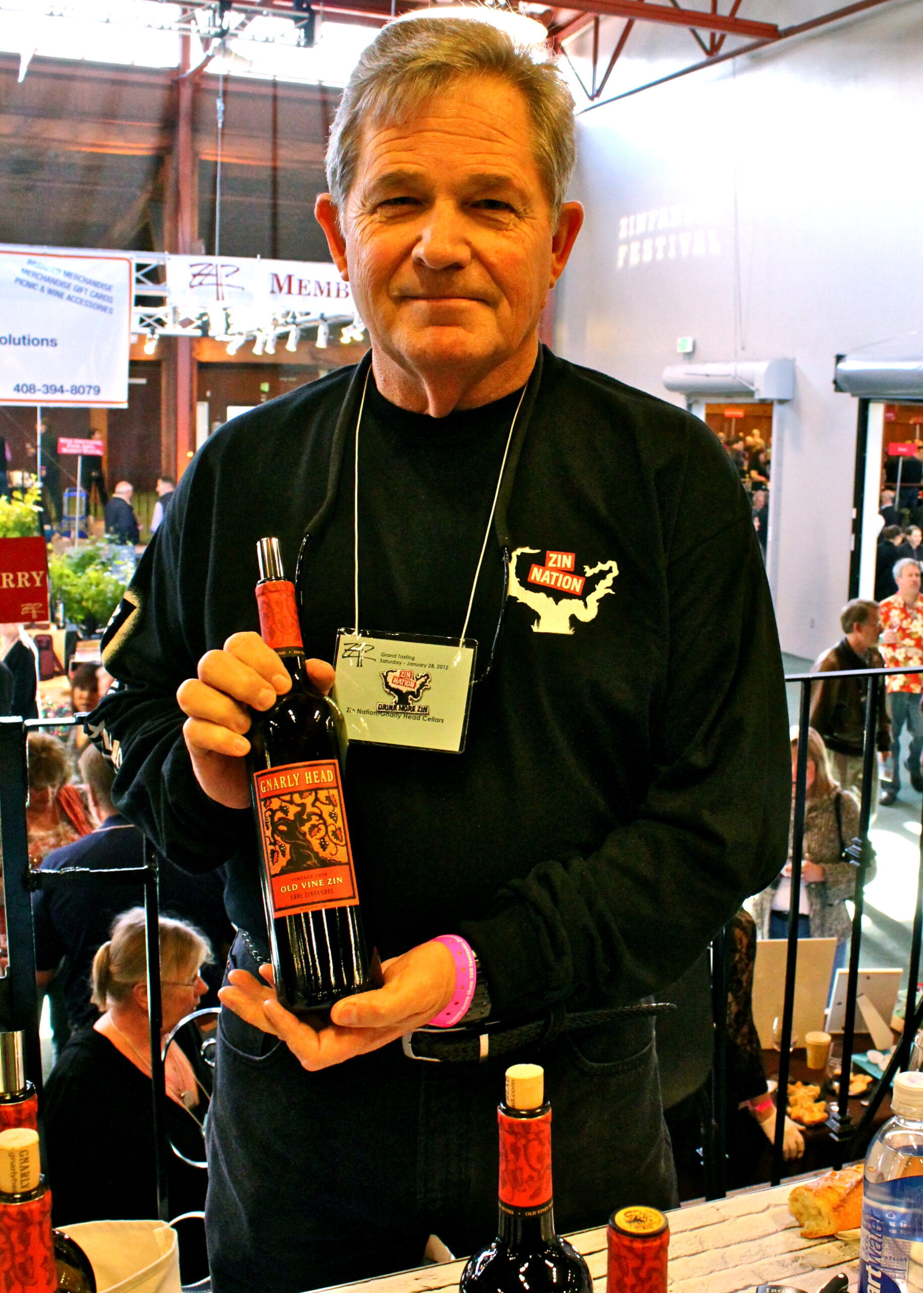 Man holding a bottle of wine at a wine event, with a busy indoor setting in the background.