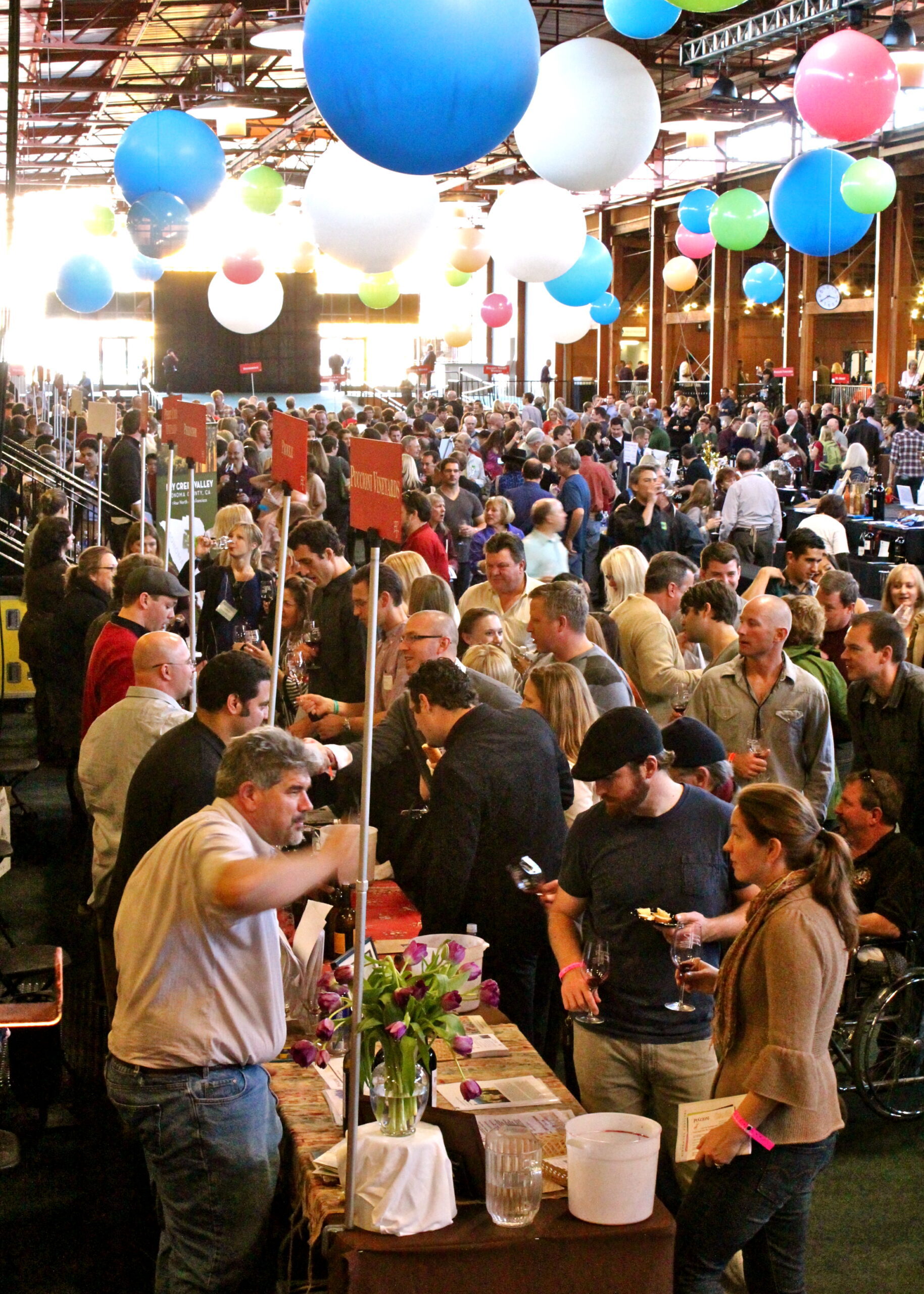 Crowd at Lodi Winegrape Commission event with colorful hanging balloons and wine tasting stations.