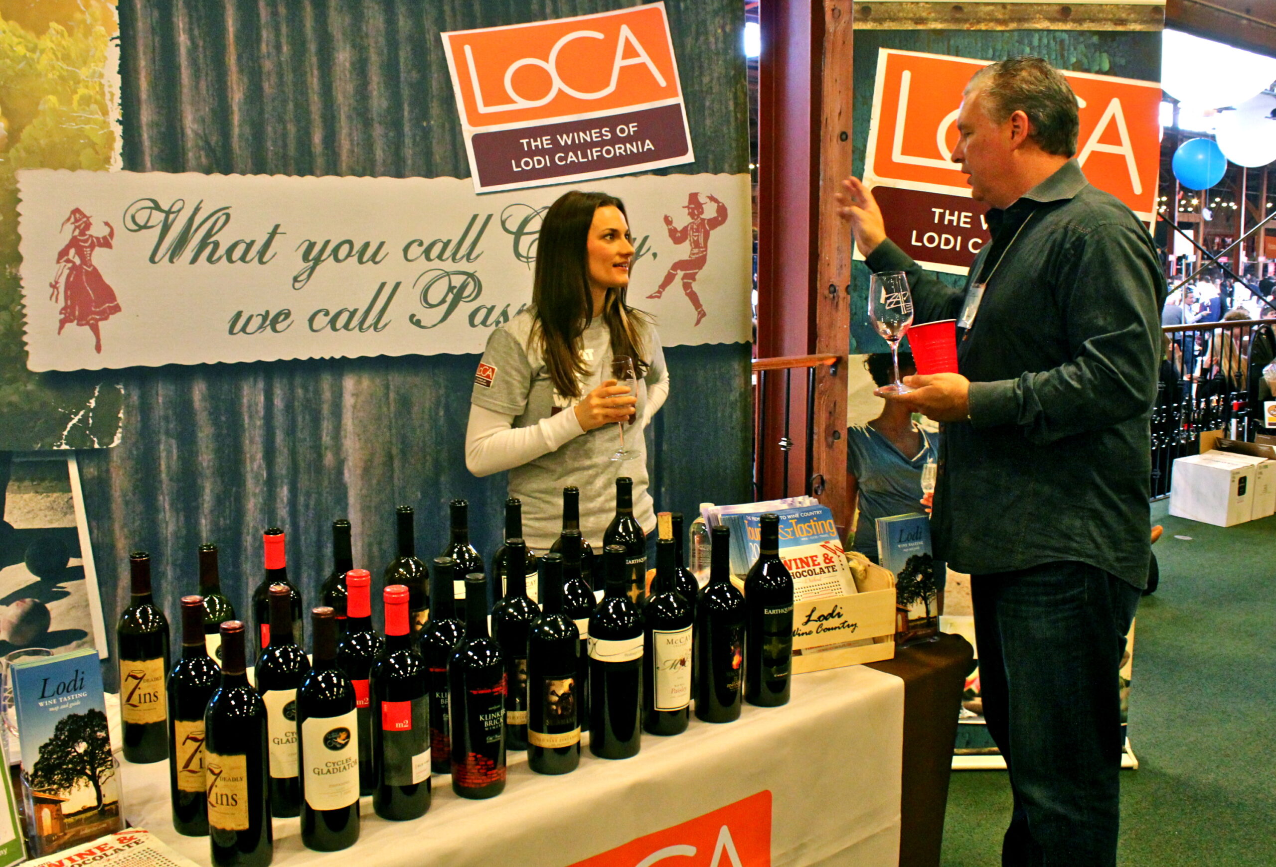 Lodi Winegrape Commission booth showcasing wines at a wine event with a representative and visitor.