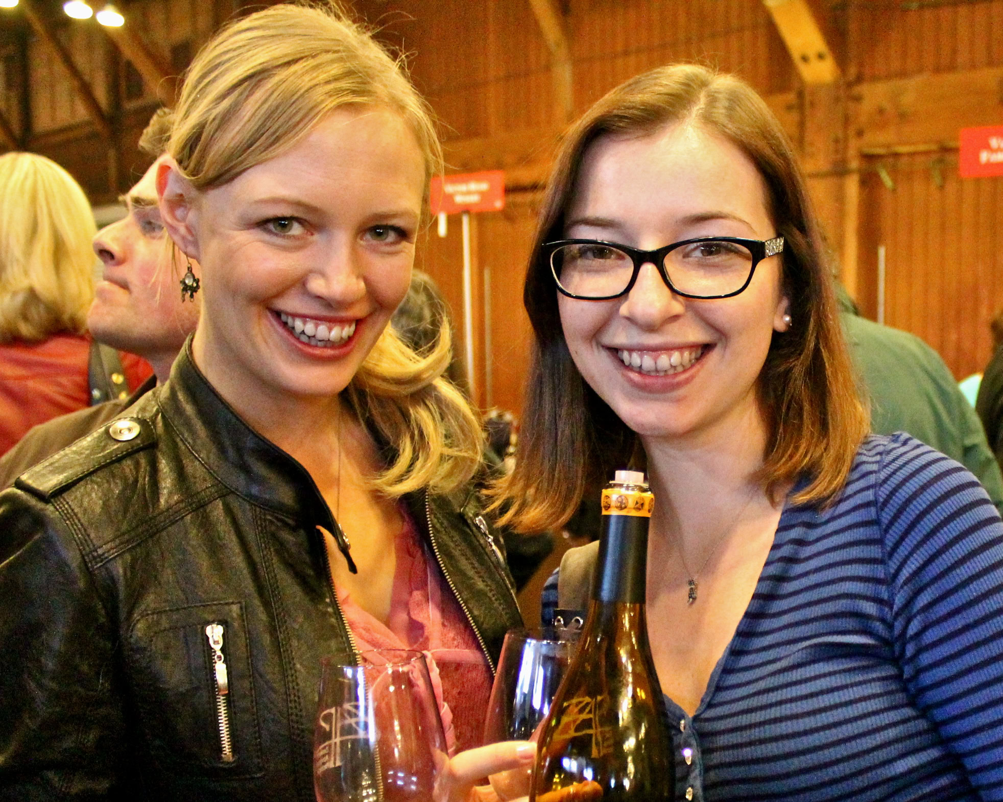 Two smiling women at a wine tasting event, holding wine glasses and a bottle, in a rustic indoor setting.