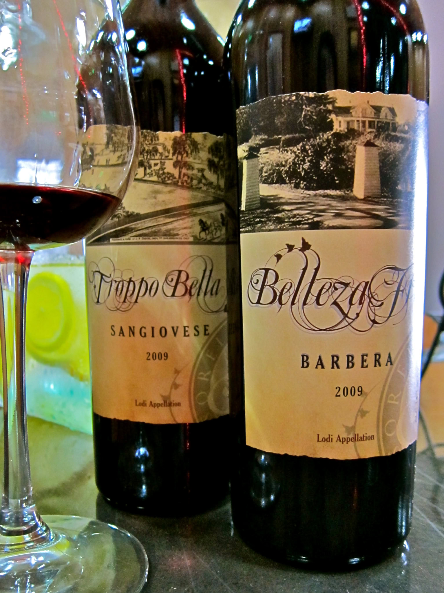 Two bottles of Lodi wine, Sangiovese and Barbera, with a glass of red wine in front, on a dark surface.