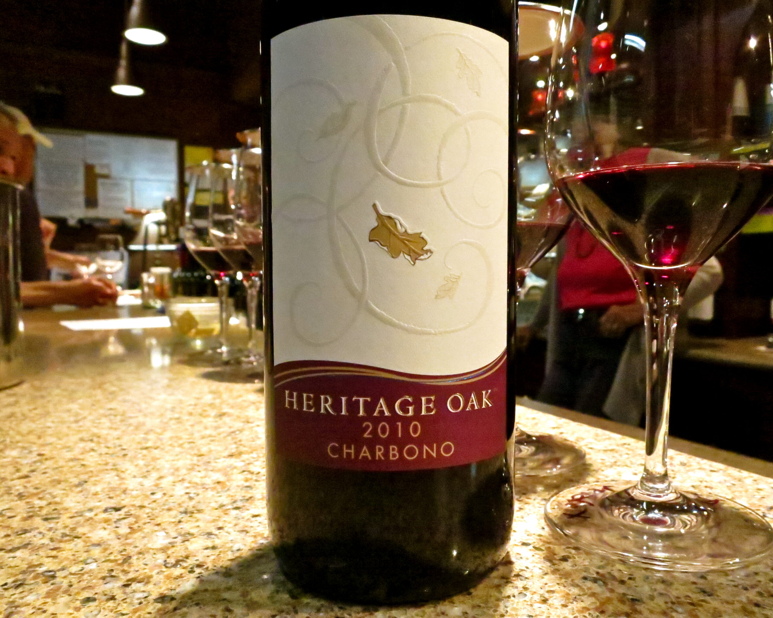 Bottle of Heritage Oak 2010 Charbono wine with two glasses of red wine on a bar counter.