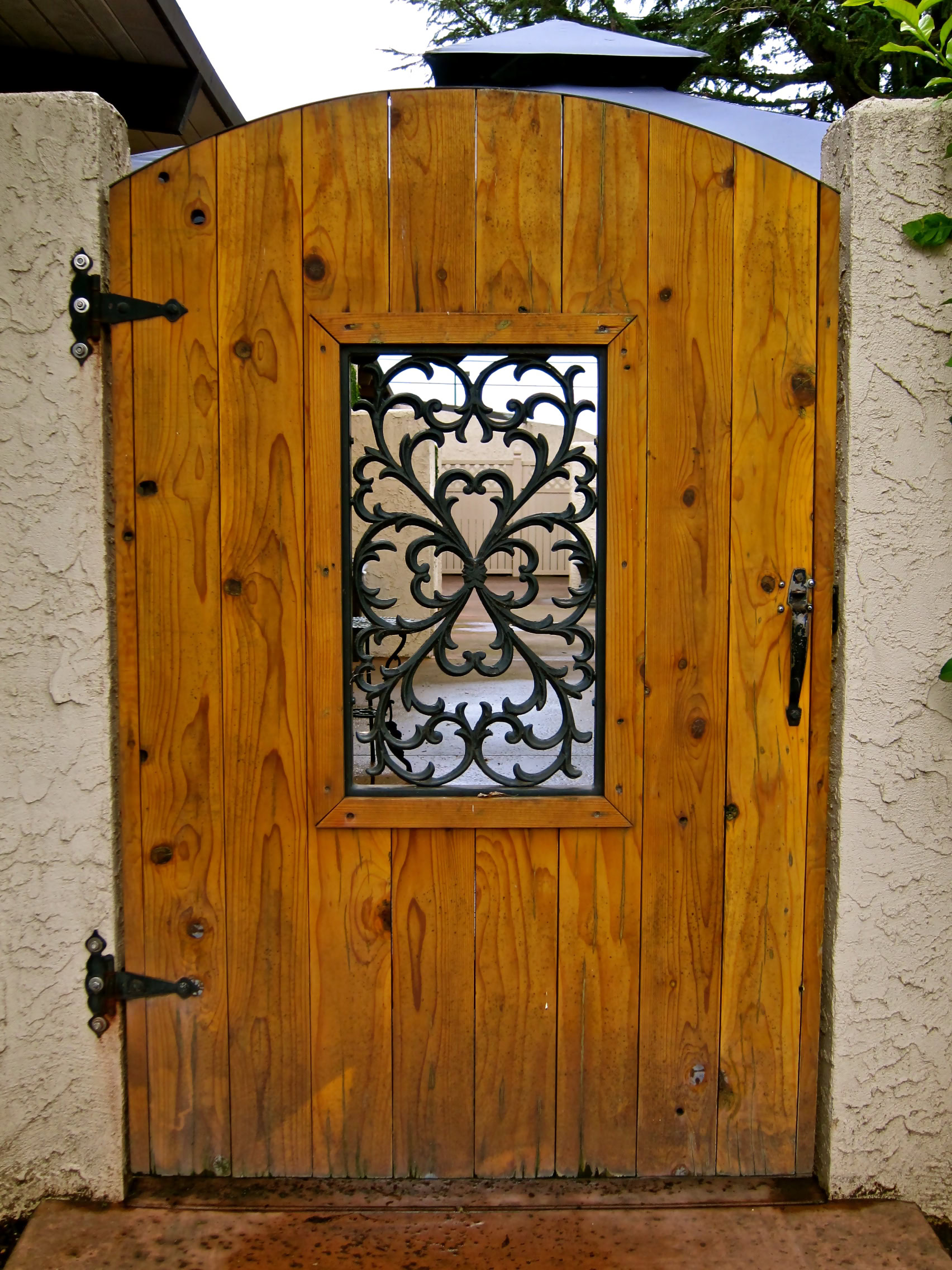 Decorative wooden gate with iron scrollwork and latch, set between stucco walls, in a garden setting.