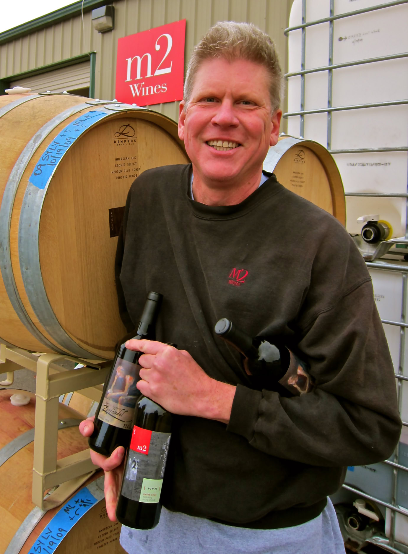 Happy winemaker holding a bottle of wine in a winery with wine barrels in the background.