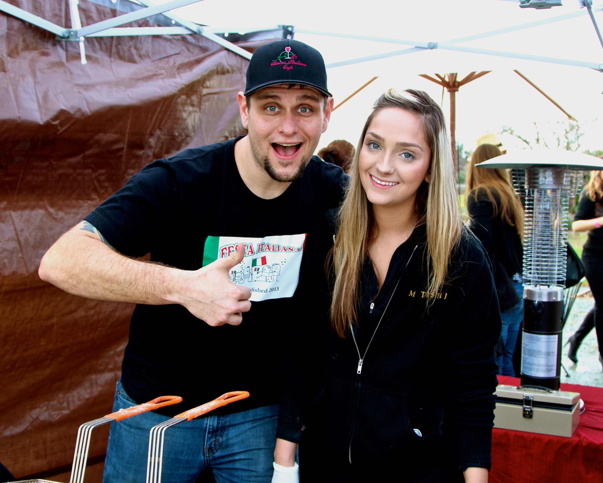 Two smiling people at a Lodi wine event, one giving a thumbs-up, with tents and other attendees in the background.