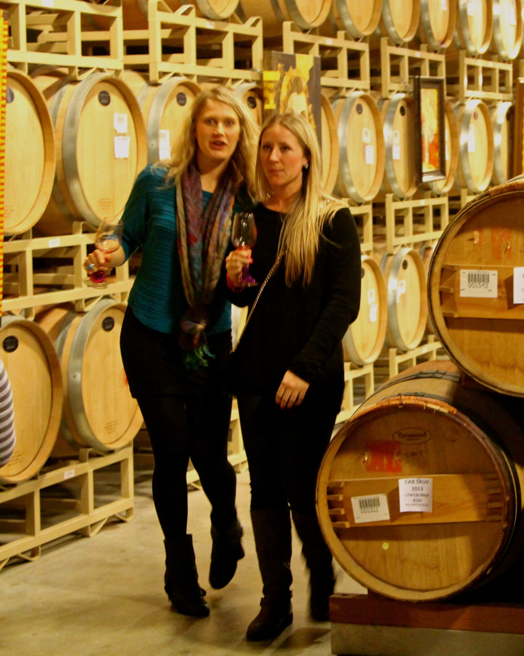 Two women tasting wine in a wine cellar with barrels in the background.