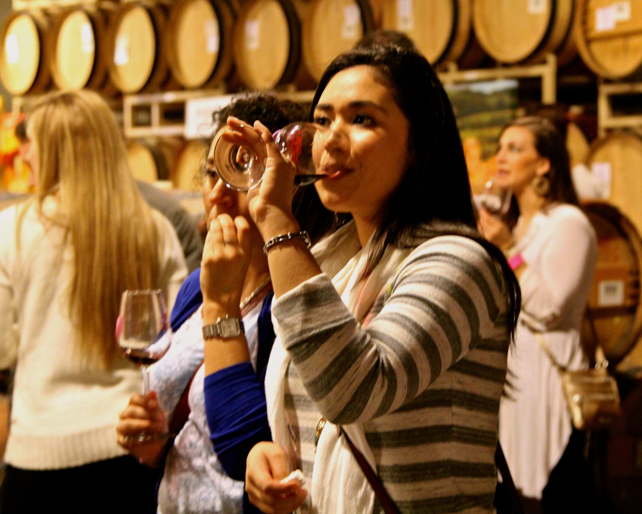 Woman tasting wine at Lodi Winegrape Commission event with barrels in background.