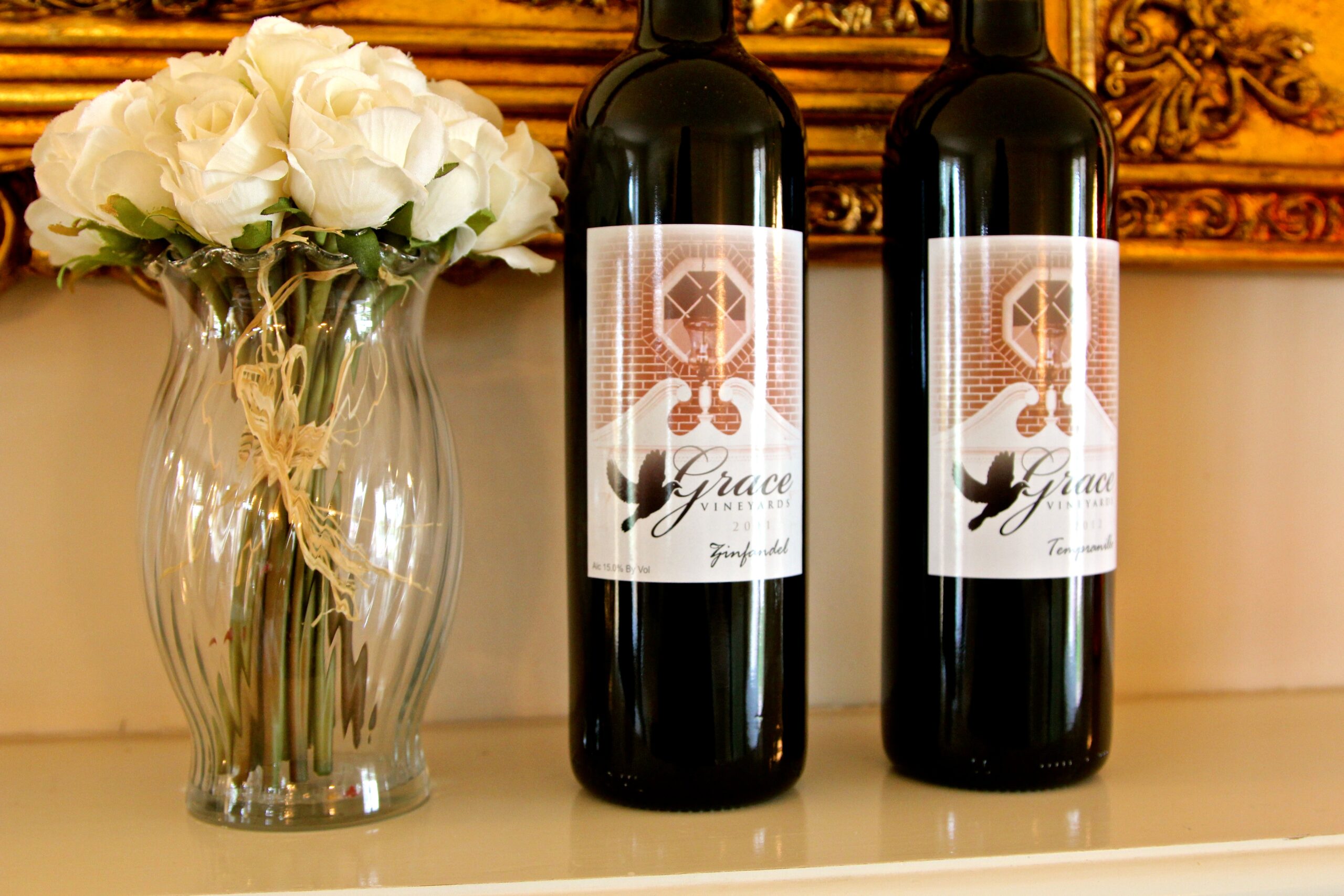 Two wine bottles with Lodi Winegrape Commission label beside a white rose bouquet in a glass vase.