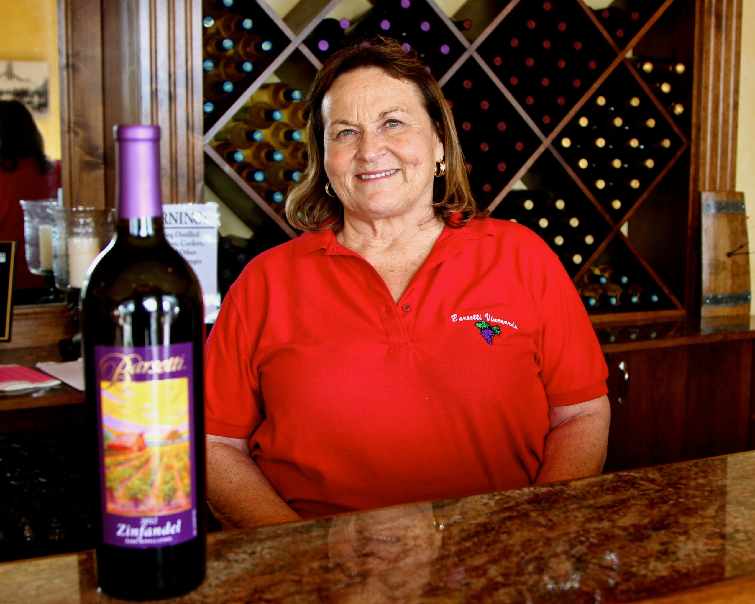 Smiling woman in red polo shirt at wine tasting bar with wine bottles and wine rack in background.