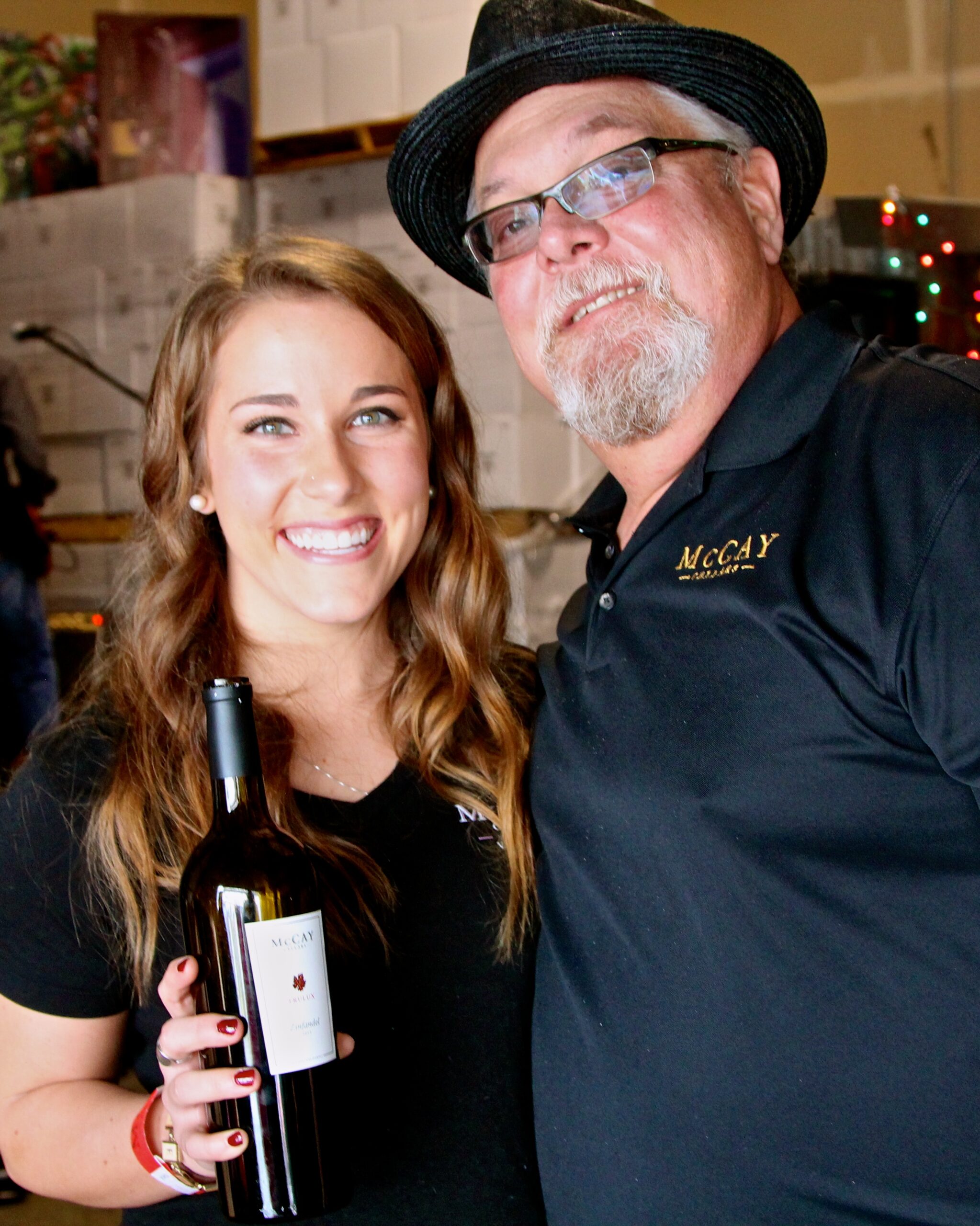 Smiling woman holding a wine bottle next to a man in a hat at a wine event.