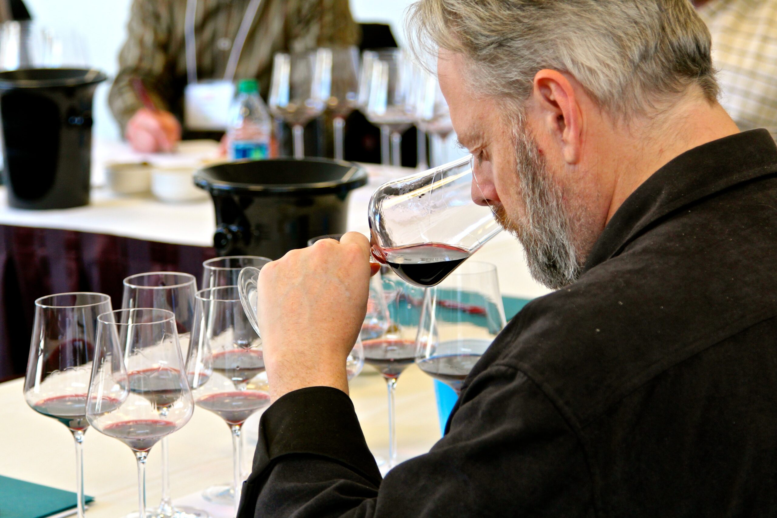 Man tasting red wine at a wine tasting event with multiple glasses and wine bottles in the background.