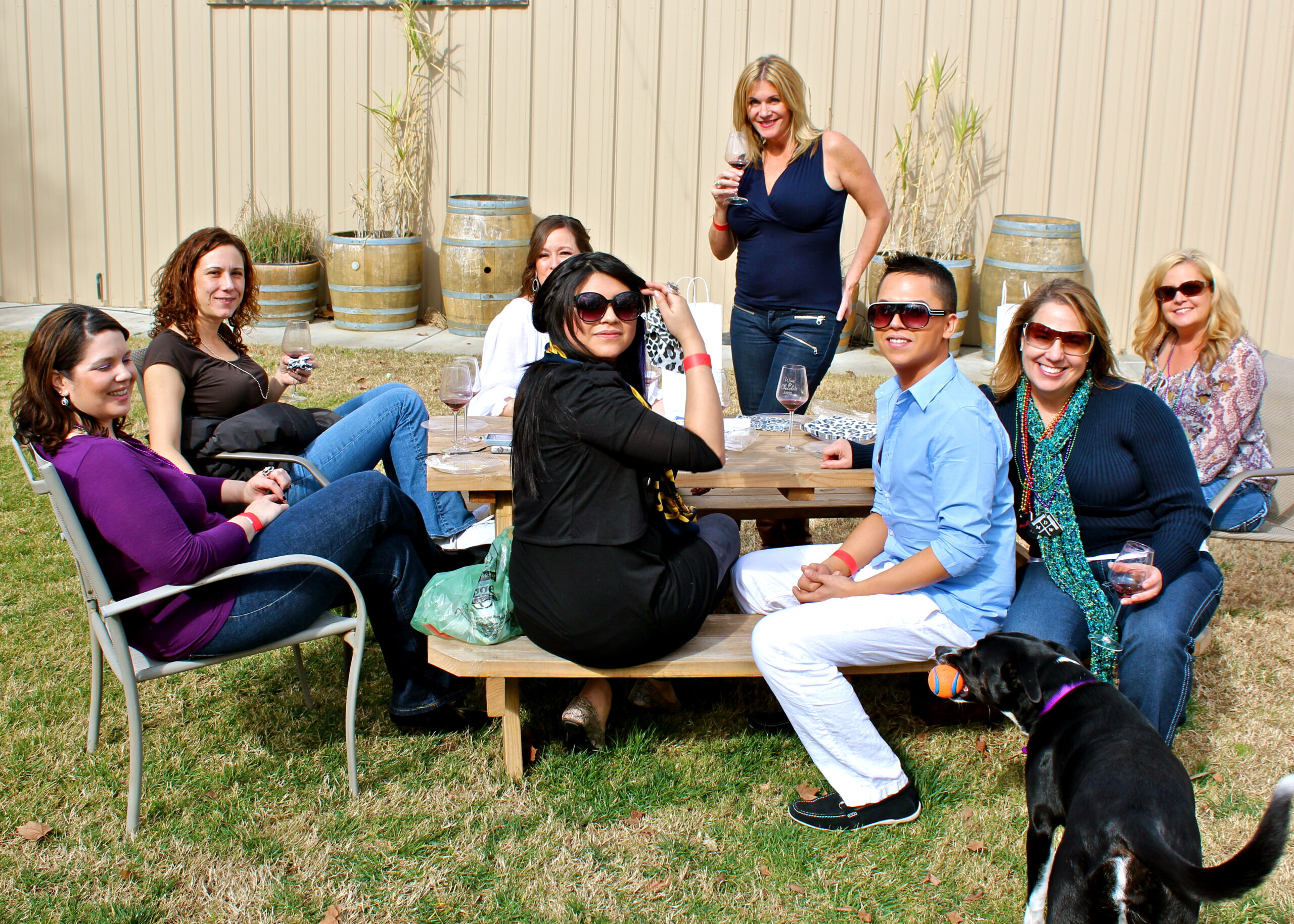 Group of people enjoying wine tasting outdoors in a garden setting with wine barrels and a dog present.