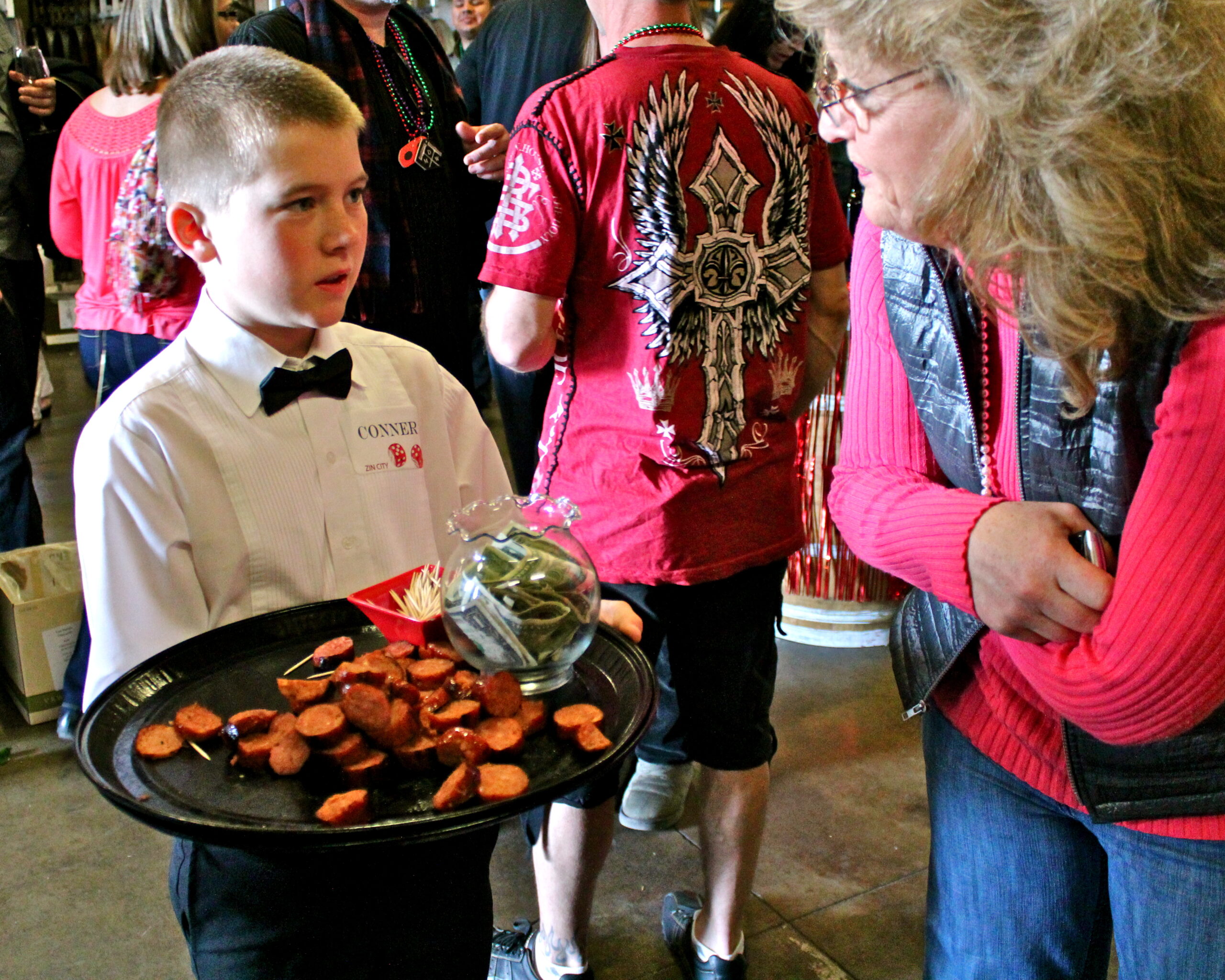 Young boy serving wine and snacks at a Lodi wine event with attendees in the background.