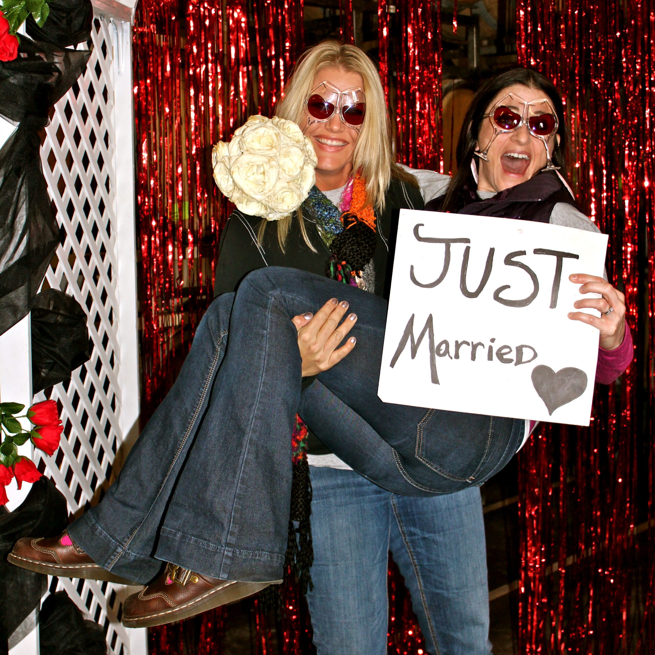 Two women celebrating marriage, holding a 'Just Married' sign, smiling, with a festive red backdrop.