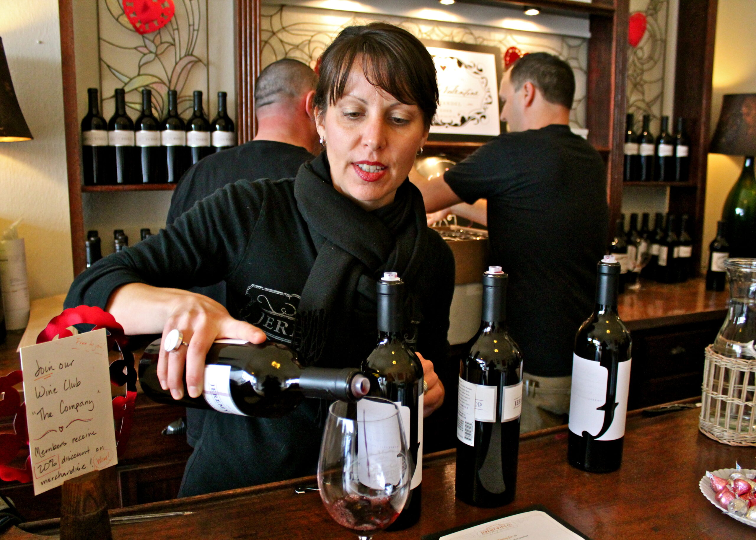 Woman pouring red wine at a tasting event with wine bottles in the background.