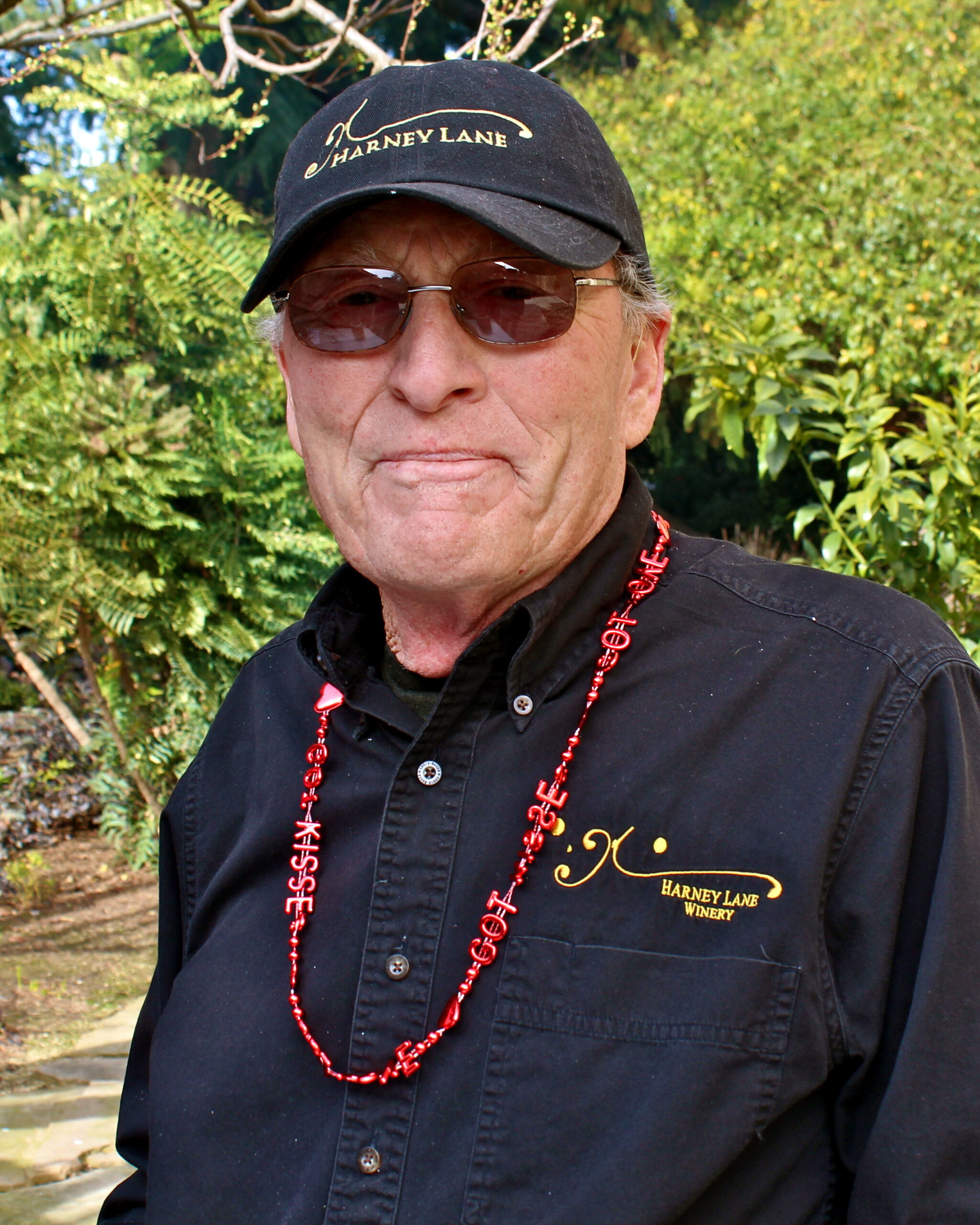 Portrait of a man wearing a black Harney Lane cap and shirt, with sunglasses, outdoors among greenery.