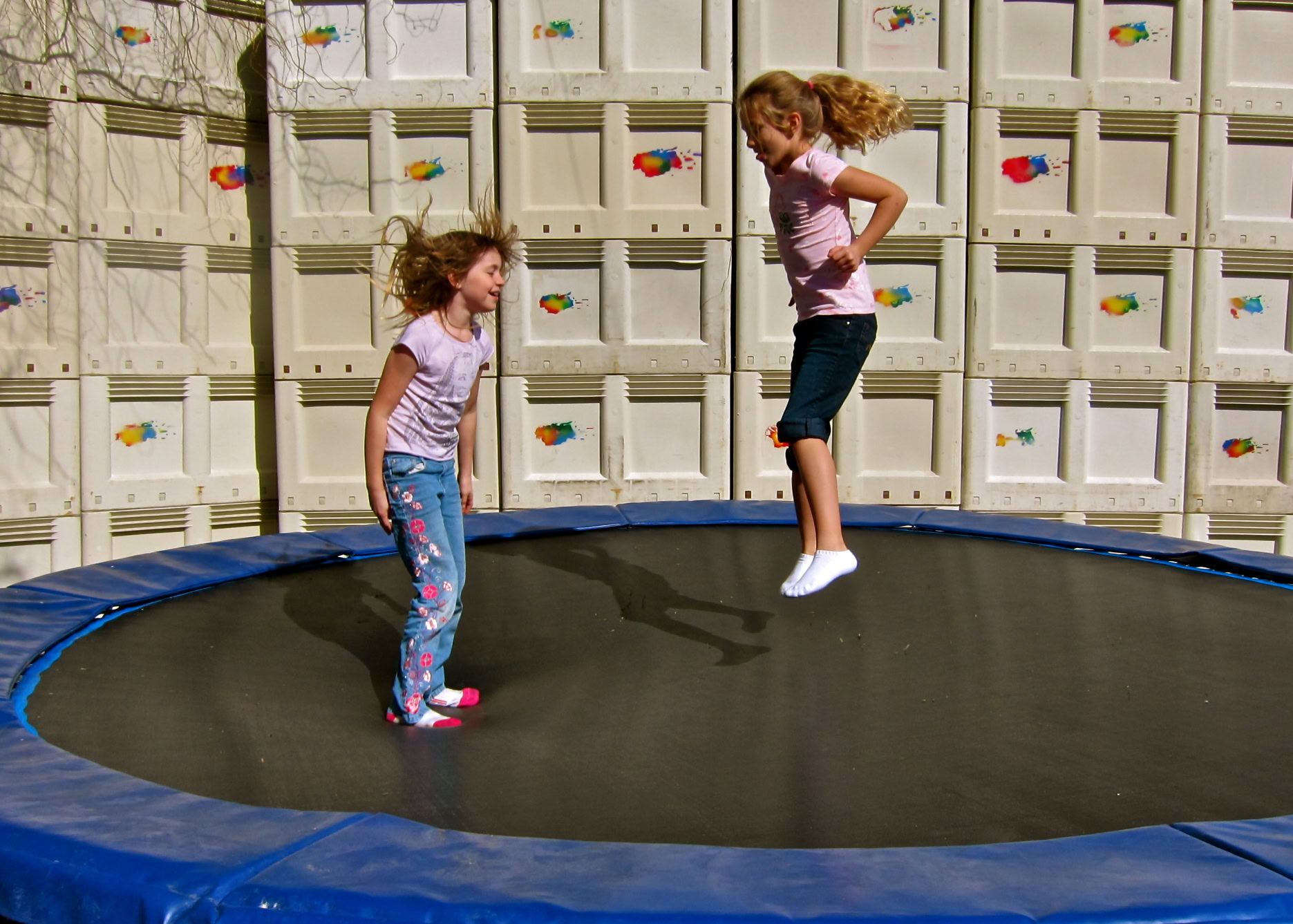 Two young girls jumping on a trampoline in a backyard with a white fence.