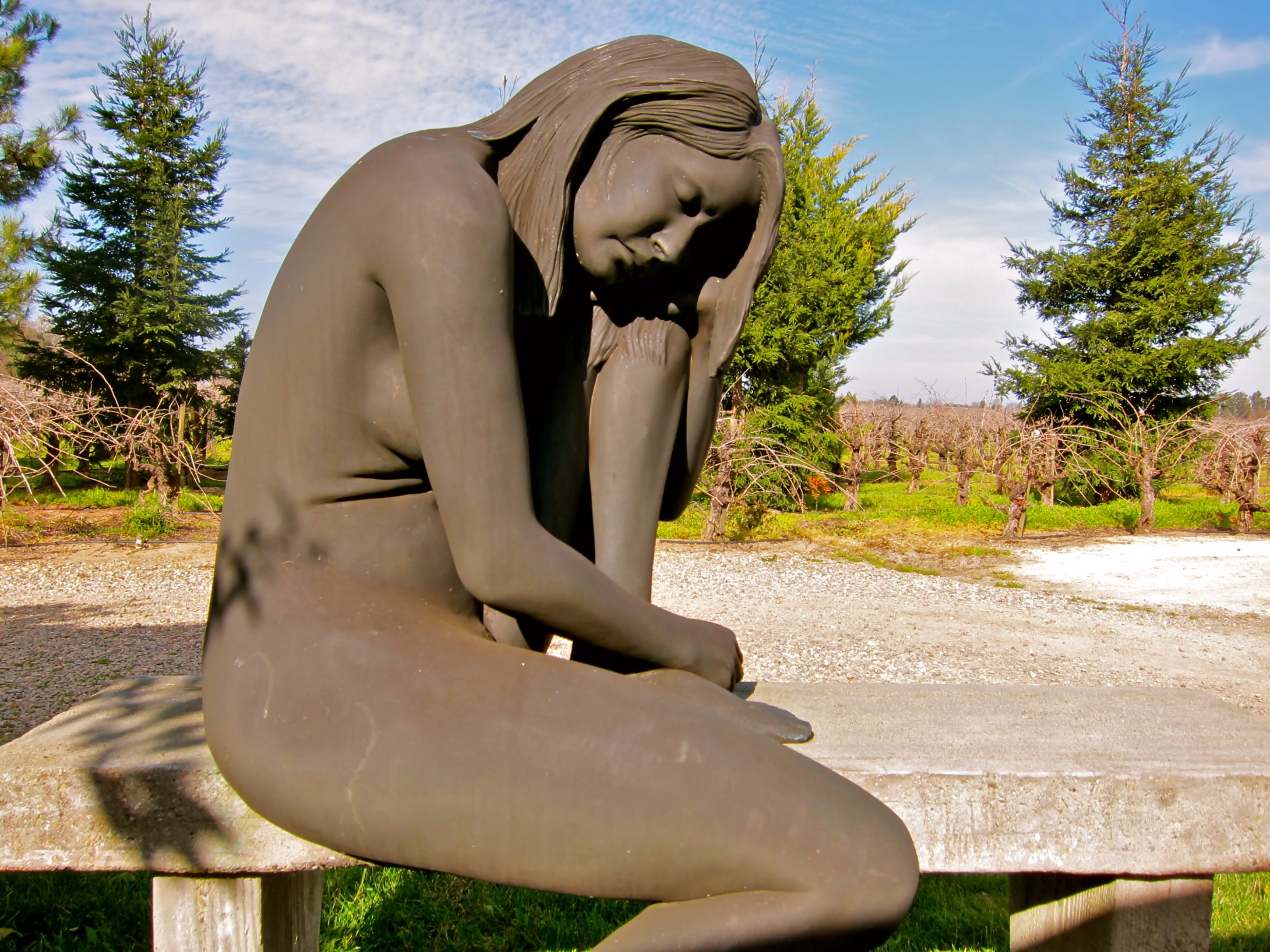 Sculpture of a contemplative woman sitting outdoors with trees and blue sky in the background.