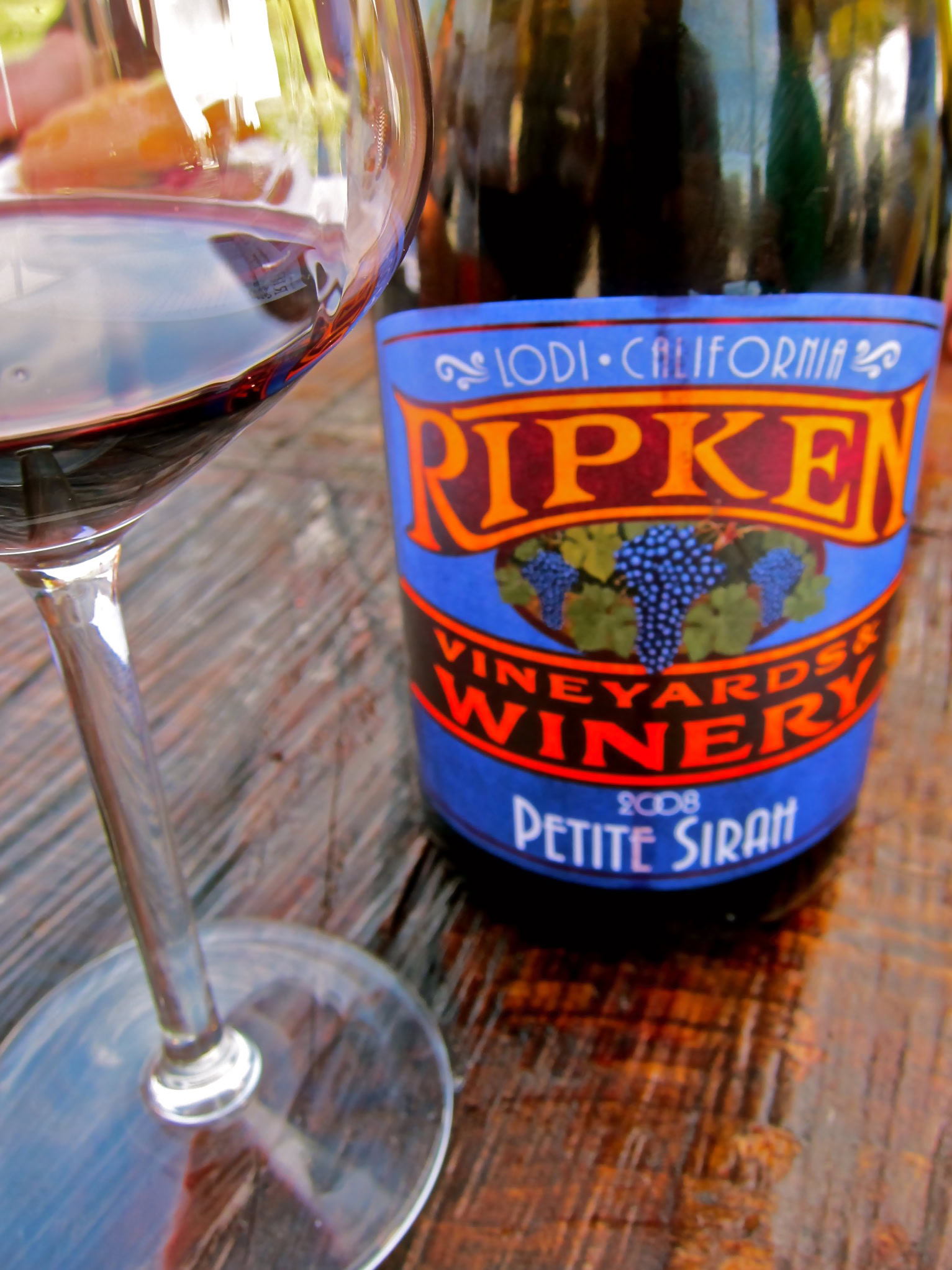 Glass of red wine with Ripken Vineyard Winery Petite Sirah bottle in background.