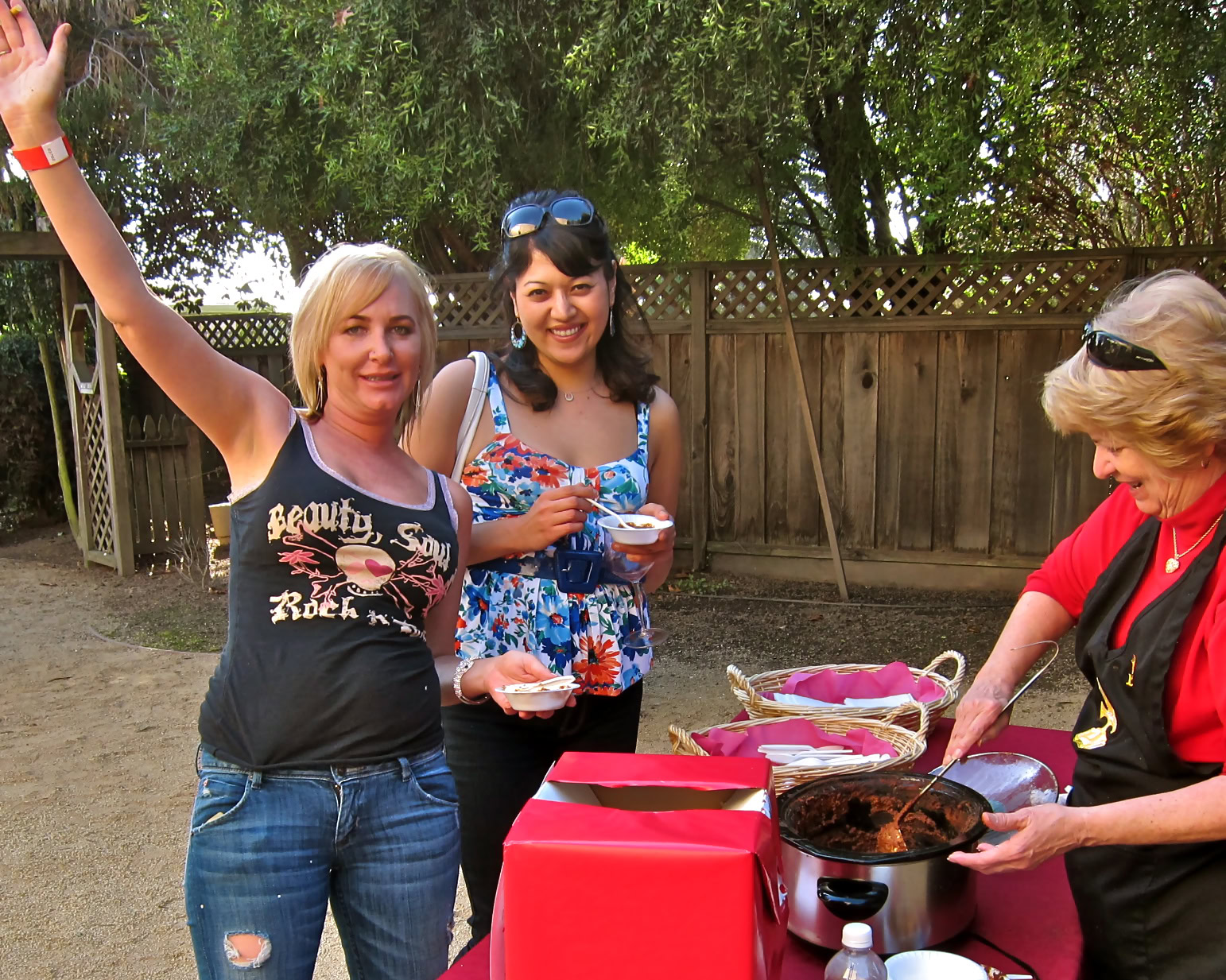 Group of women enjoying wine tasting at an outdoor event with a barbecue grill and trees in the background.