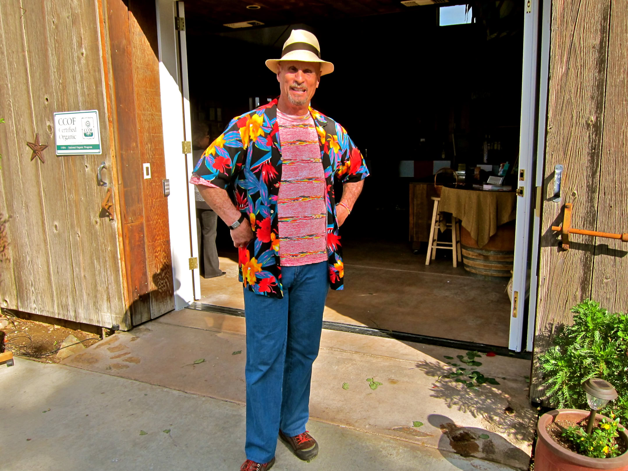 Man in colorful Hawaiian shirt and straw hat standing outside winery entrance.