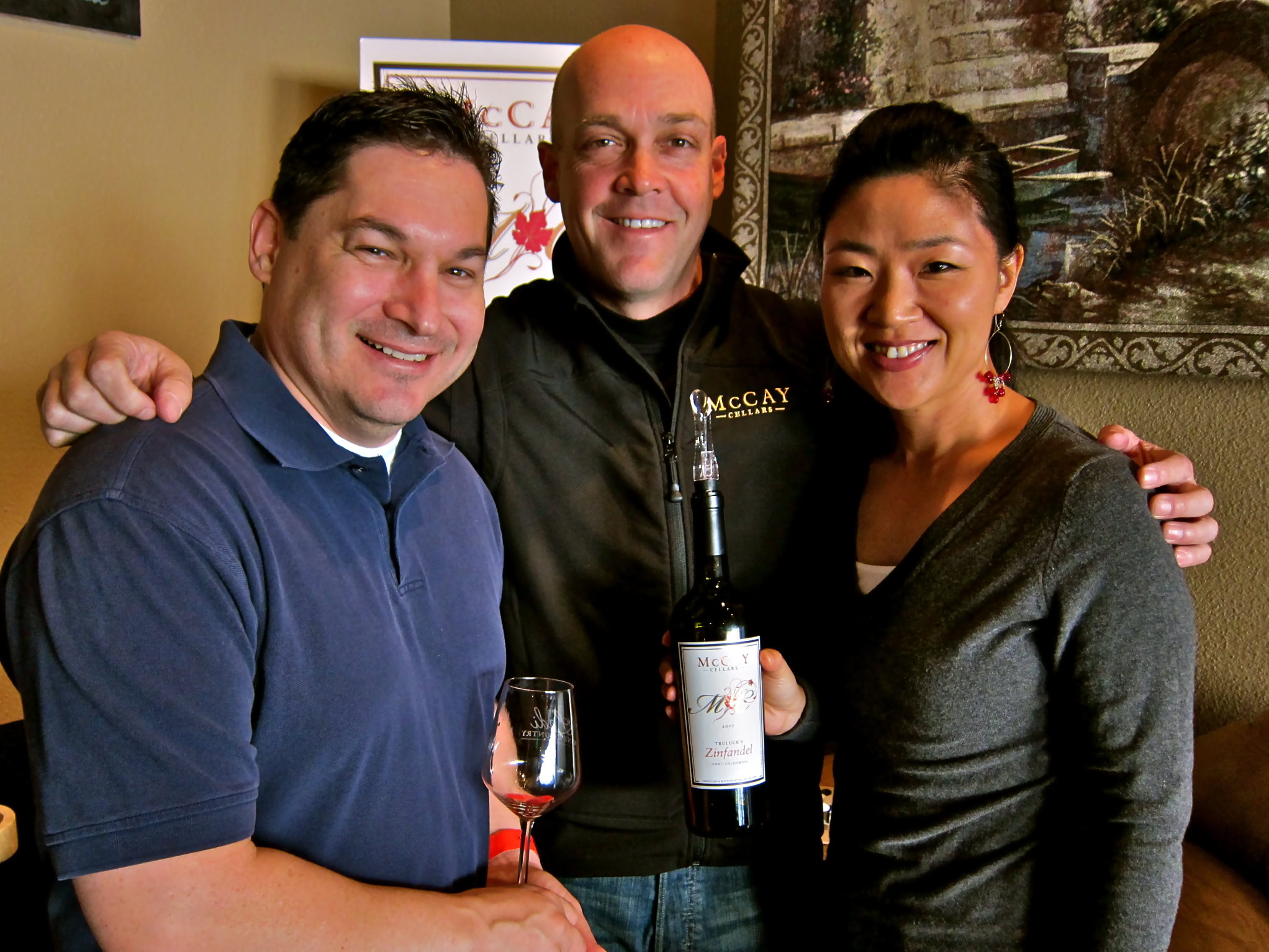 Group of three people at a wine tasting event, holding a wine bottle and glass, smiling for the camera.