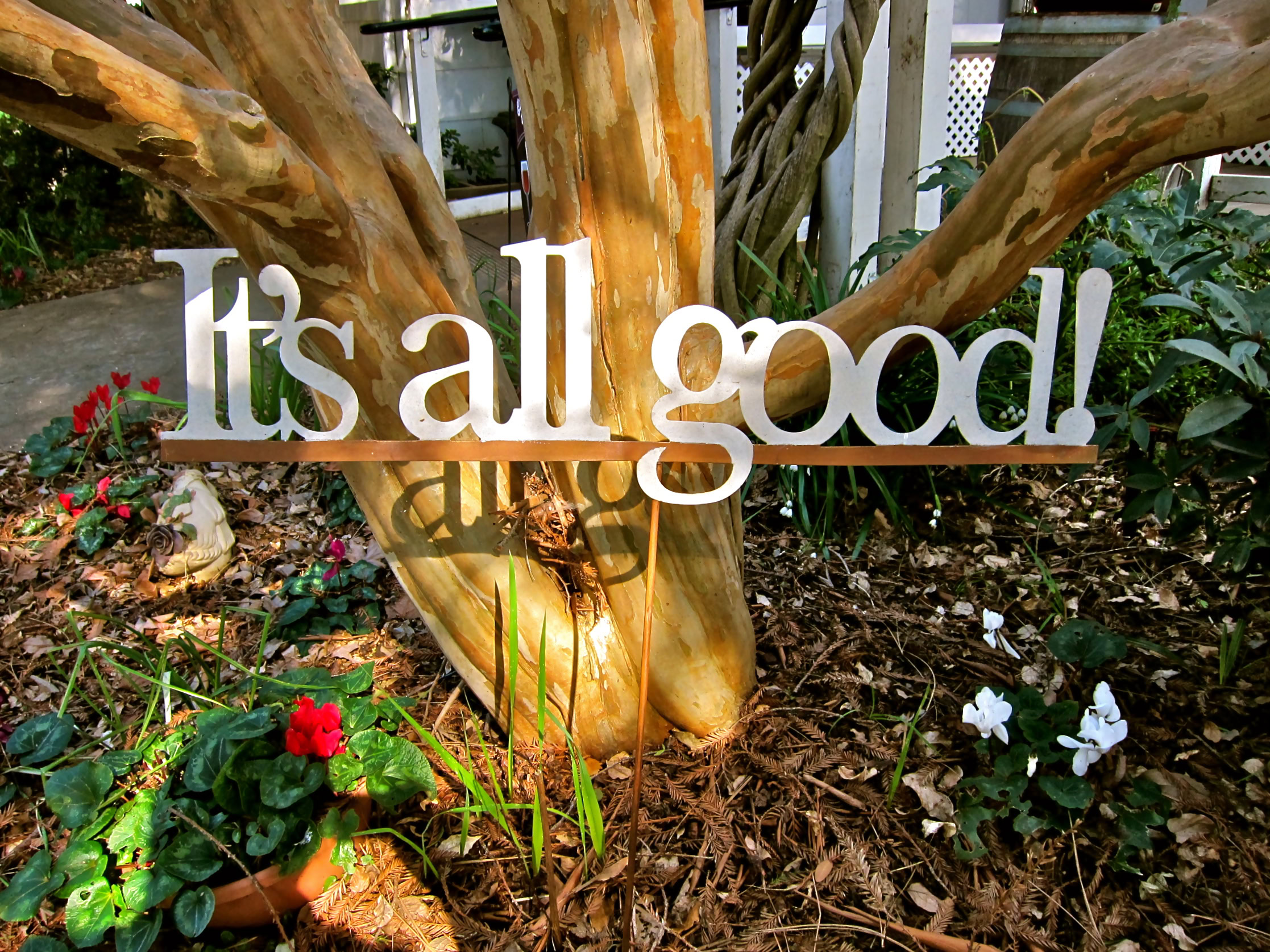 Decorative sign reading 'it's all good' in a garden with flowers and a tree trunk.