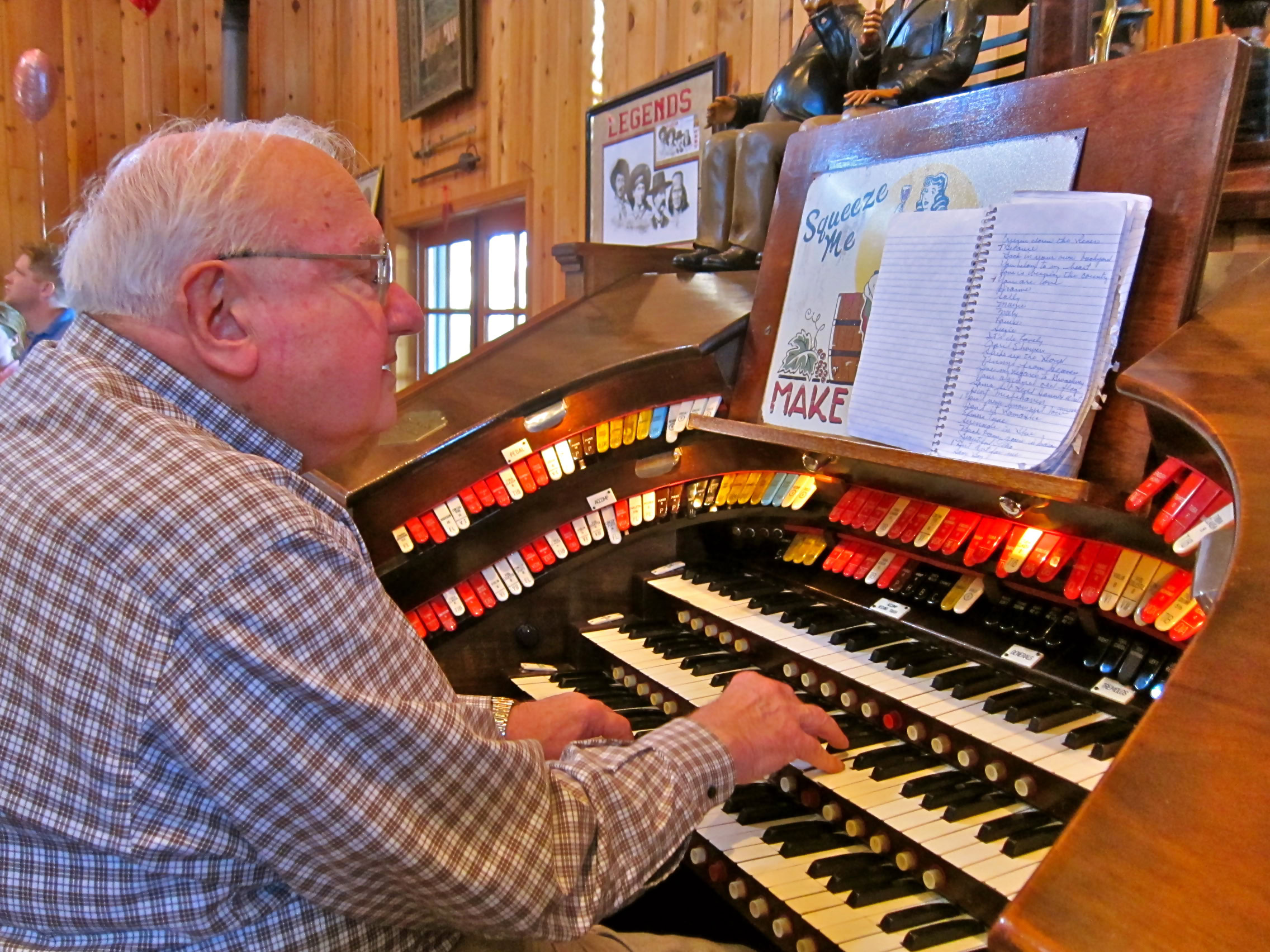 Elderly man playing a vintage pipe organ in a cozy wooden interior, surrounded by wine-themed decor and memorabilia.