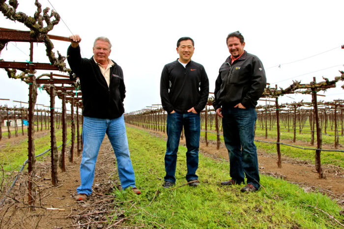 Three men standing in a vineyard, representing the Lodi Winegrape Commission, with grapevines and trellises in the background.