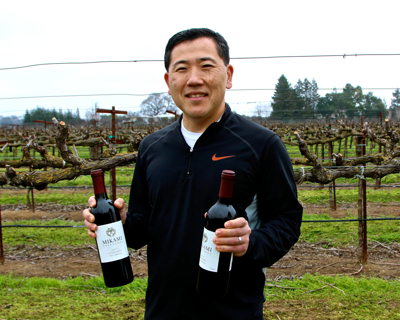Man holding two bottles of wine in a vineyard during daytime, smiling at camera.