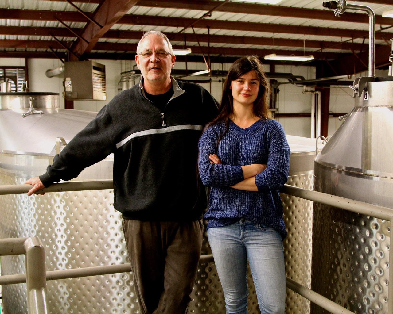 Two people standing inside a winery with large stainless steel fermentation tanks in Lodi, California.