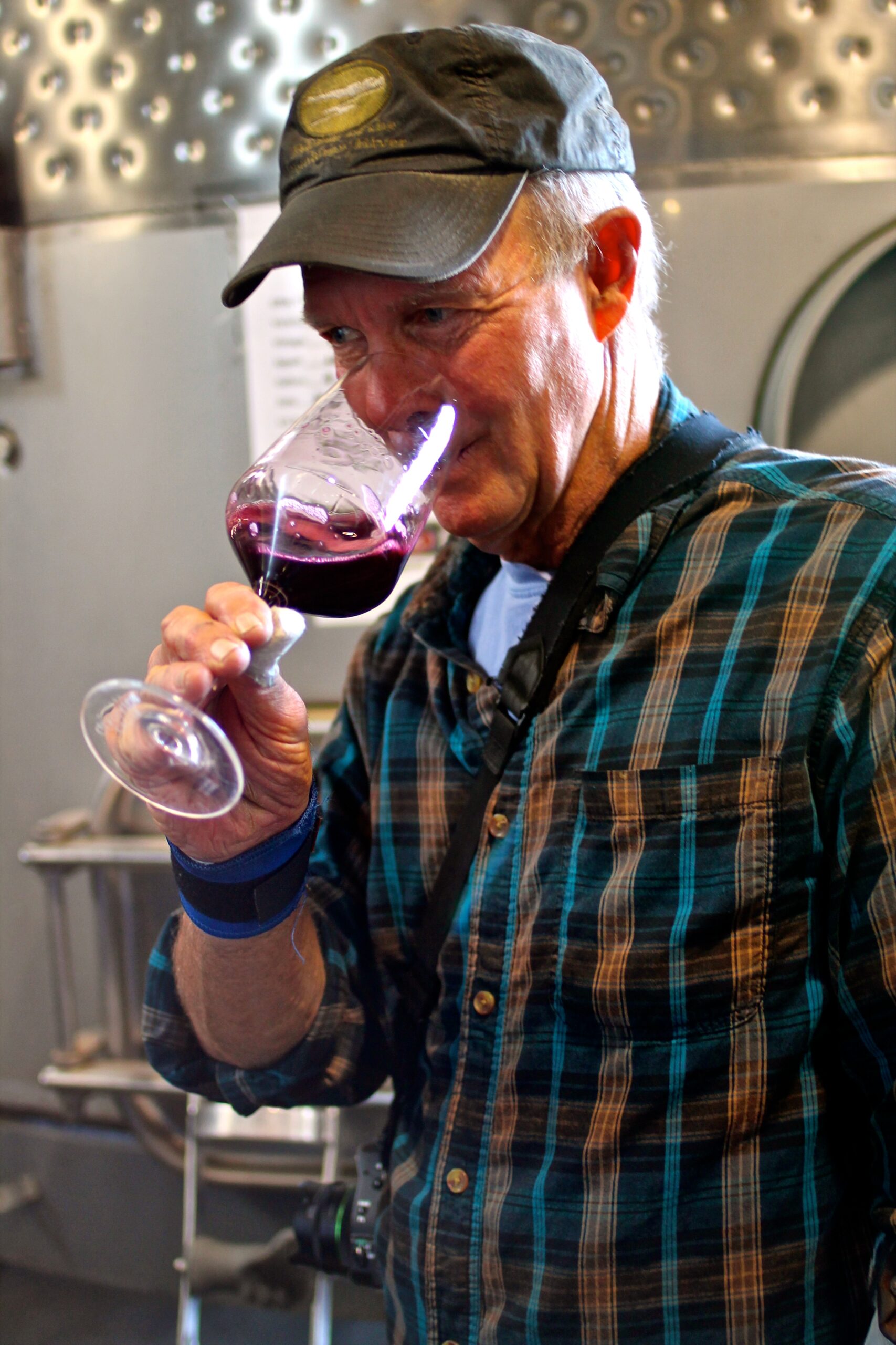 Man tasting red wine at Lodi Winegrape Commission event, wearing a cap and plaid shirt, in a winery setting.