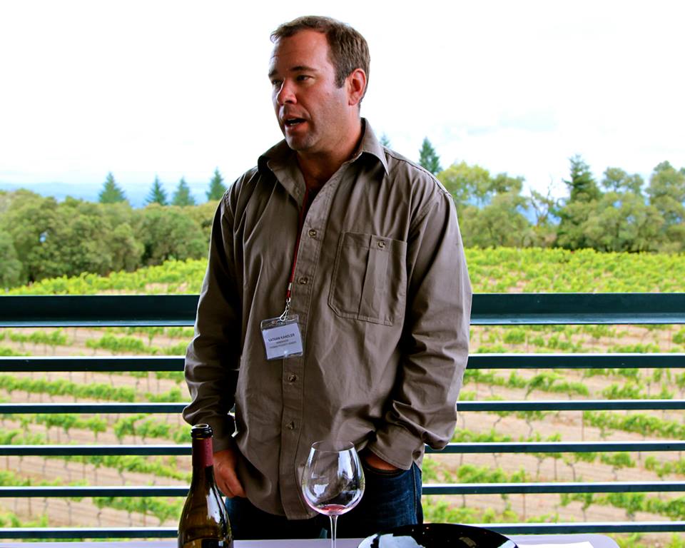 Man speaking at a wine tasting event with vineyards in the background.