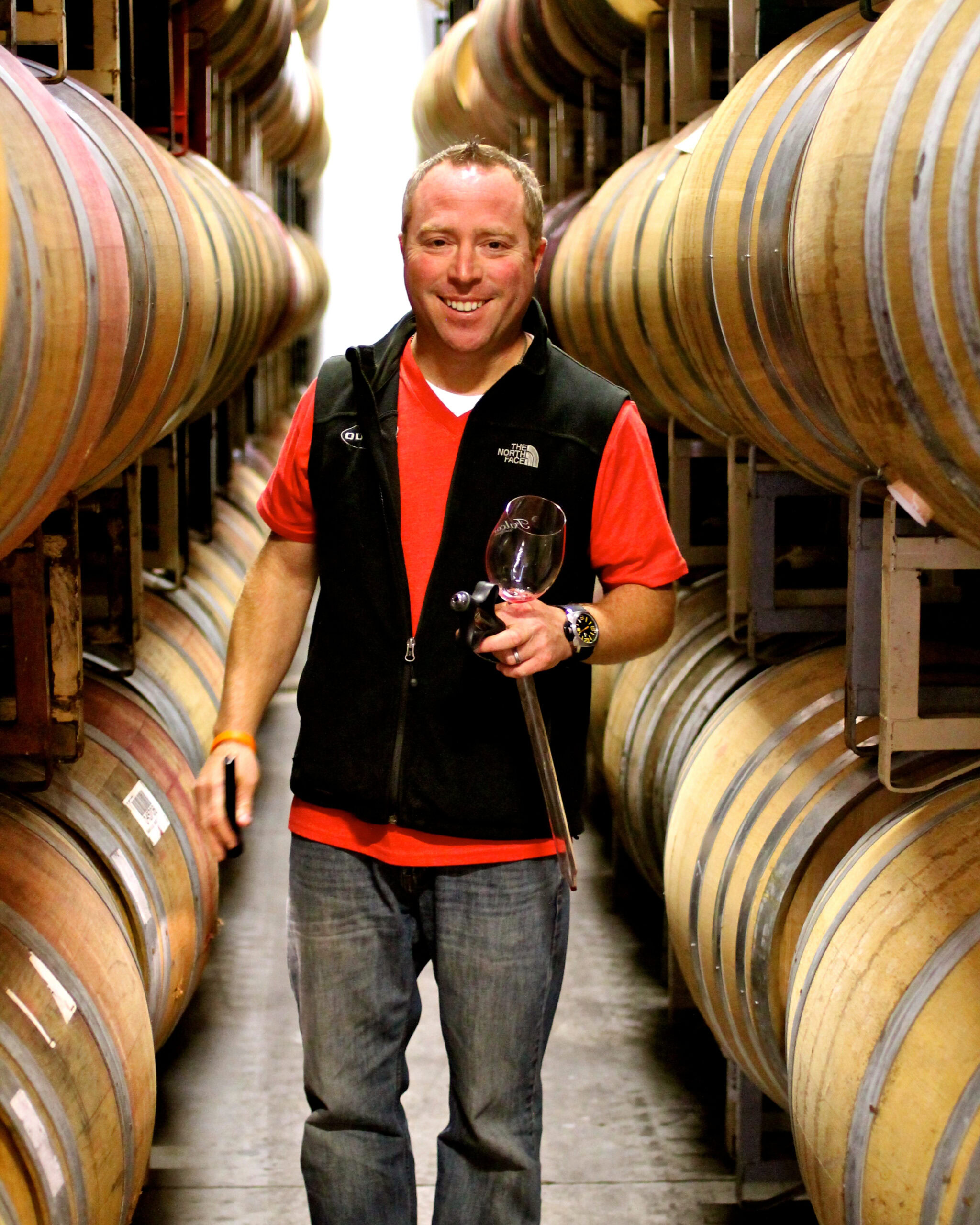 Man holding a glass of red wine in a wine cellar with barrels on both sides.