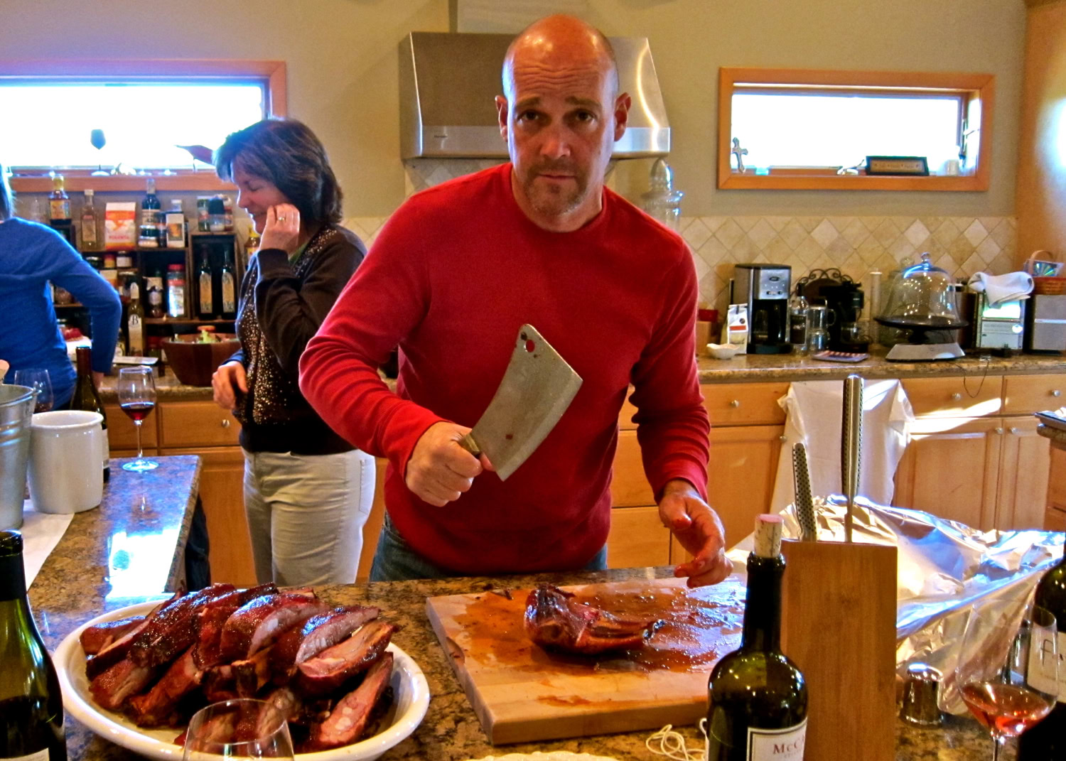 Man preparing meat at a cooking event, with wine bottles and kitchen in background.