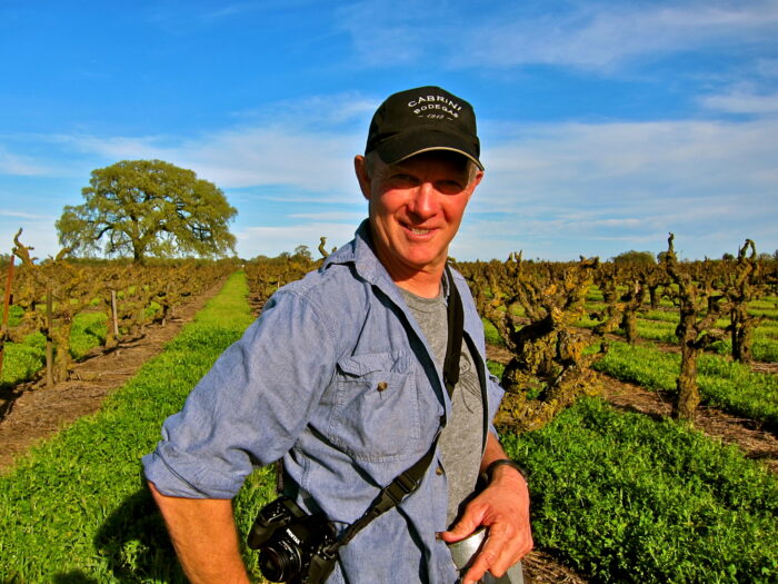 Smiling man in vineyard with rows of grapevines under blue sky, wearing a cap and camera, representing Lodi wine industry.